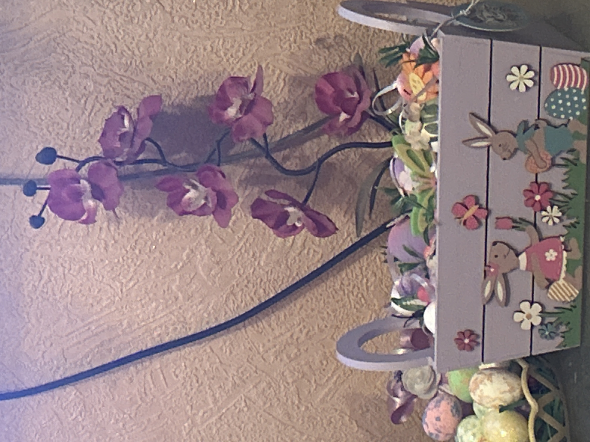 The image presents a close-up, indoor scene with a textured, light-tan wall in the background. A dark, seemingly synthetic, vine with purple flowers is attached to the wall, arching from left to right. To the right, a decorated Easter-themed object is prominent. It features a light purple, plank-like structure adorned with cut-out images of rabbits, flowers, and Easter eggs. Ribbons and additional flowers are attached around the object. Below, faux Easter eggs in a variety of colors are visible, partially contained within a woven basket. The scene suggests a decorative display, likely related to Easter celebrations. There is no indication of location in Sofia, Bulgaria.