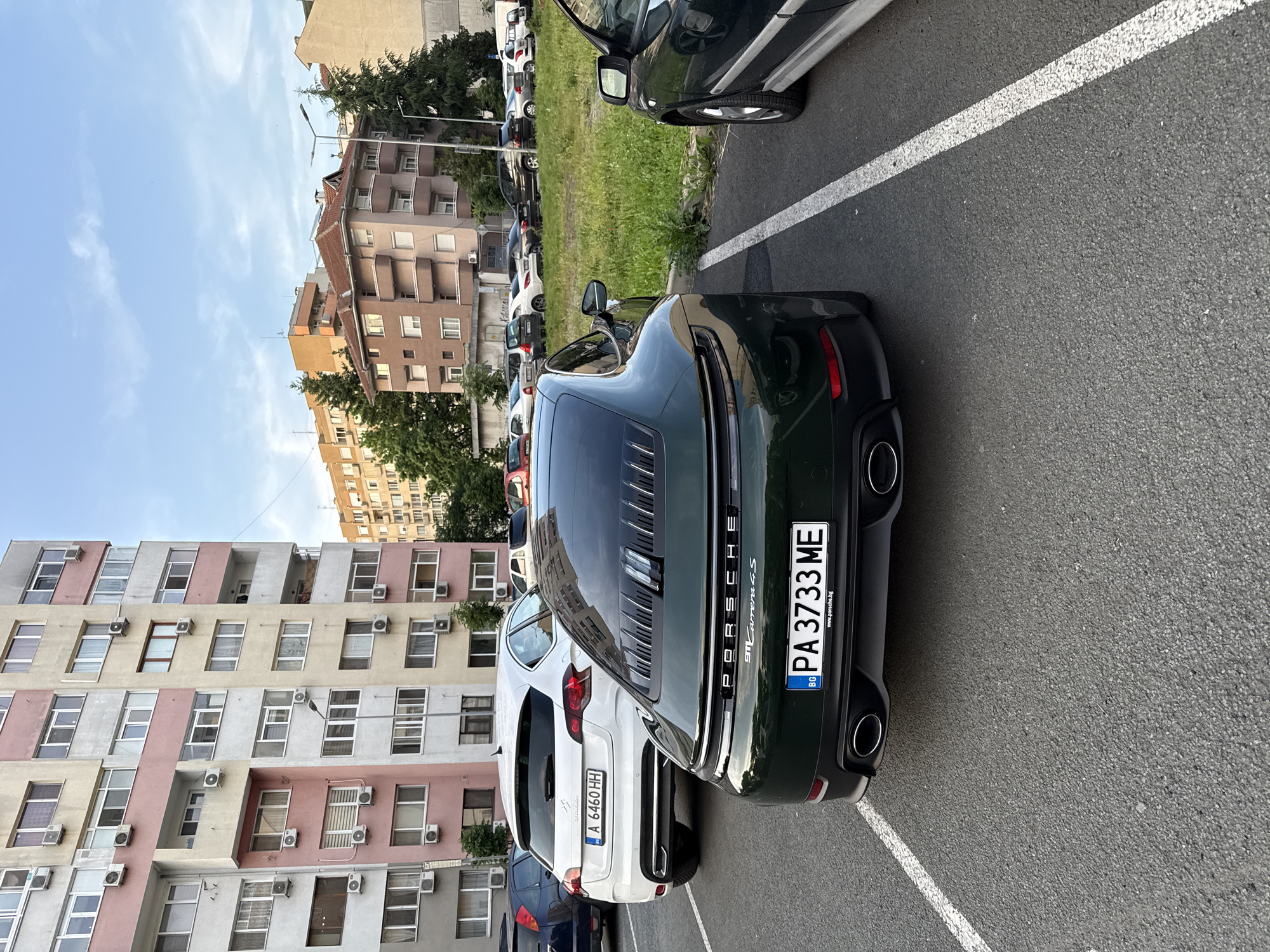 The image captures the rear of a dark green Porsche 911 Carrera S parked in a lot, positioned next to a white Citroen C-Crosser. The Porsche's license plate reads "PA 3733 ME", and the Citroen's license plate reads "A 6460 HH", both being Bulgarian license plates. The vehicles are parked in marked spaces, with the Porsche angled slightly within its space. Behind the cars, there are several multi-story residential buildings with varying architectural styles and colors. A grassy area is visible behind the parked vehicles. The sky is light blue and relatively clear. This is likely in the city of Burgas, Bulgaria.
