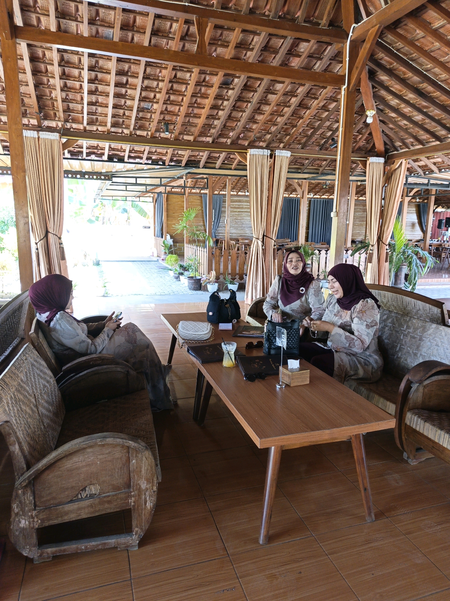 The image depicts an indoor seating area of a restaurant or cafe in Jonggrangan, Indonesia, featuring traditional Javanese architecture with a high, wooden-beamed roof tiled with terracotta. Three women wearing headscarves and modest clothing are present. One woman is seated on a woven rattan bench on the left, looking at her phone. Two other women are seated on a longer, upholstered bench on the right, facing each other and engaged in conversation. A low, wooden coffee table is situated between them, with drinks, menus, and handbags on its surface. The floor is tiled with brown ceramic squares. The space is open on the right side, revealing an outdoor area with potted plants and a white fence. The overall ambiance suggests a relaxed, casual setting.