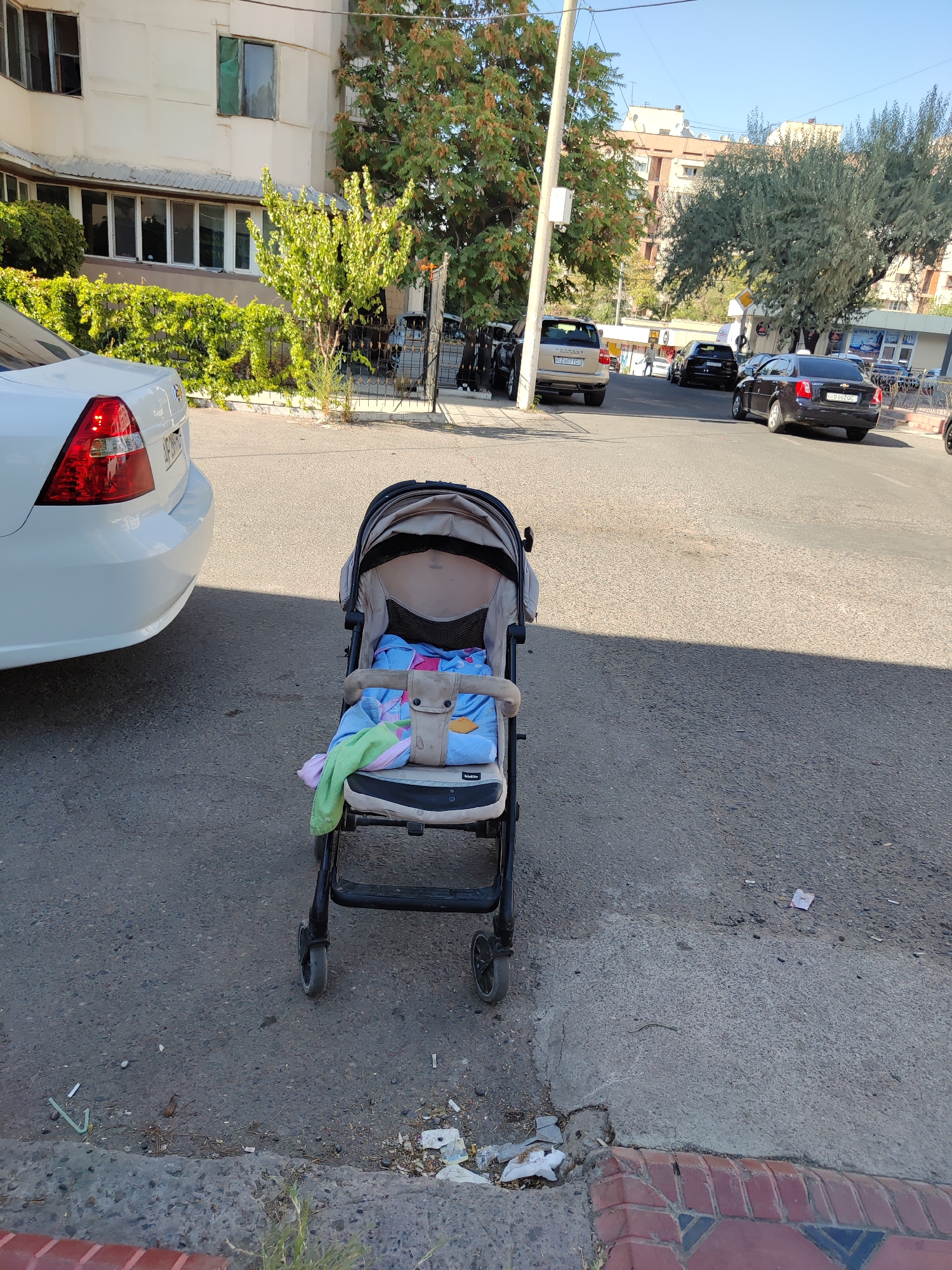 The image depicts a street scene in Tashkent, Uzbekistan. In the foreground, a stroller is parked on the asphalt. The stroller is light-colored and appears to have a blanket or some fabric inside. To the left of the stroller is the rear of a white car. In the background, there are trees, apartment buildings, and several cars parked or driving on the street. The sky is clear and blue. The scene suggests a typical urban environment.