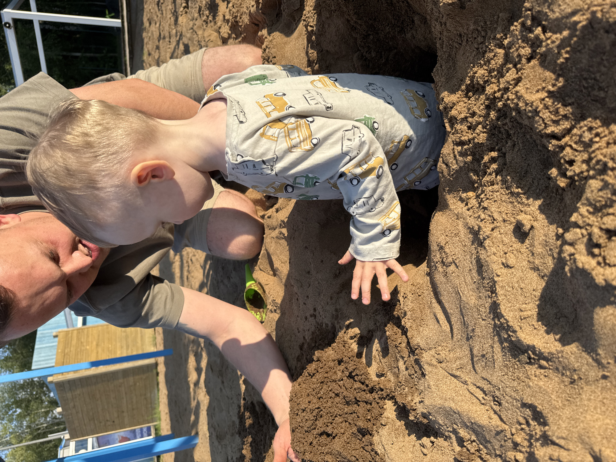 A man and a young boy are playing in a sandbox on a sunny day. The boy, wearing a long-sleeved shirt with a pattern of yellow and green vehicles, is partially submerged in a hole he has dug in the sand. His hand is visible reaching out from the hole. The man, wearing a grey t-shirt and beige shorts, is kneeling beside the boy, also digging in the sand with his hands. A green plastic shovel lies in the sand to the left. In the background, there is a wooden structure with blue accents and some greenery, suggesting an outdoor playground or park. The lighting indicates it is daytime, with the sun casting shadows. The overall mood appears to be one of joyful outdoor play. The location is specified as Chaikovsky, Russia.
