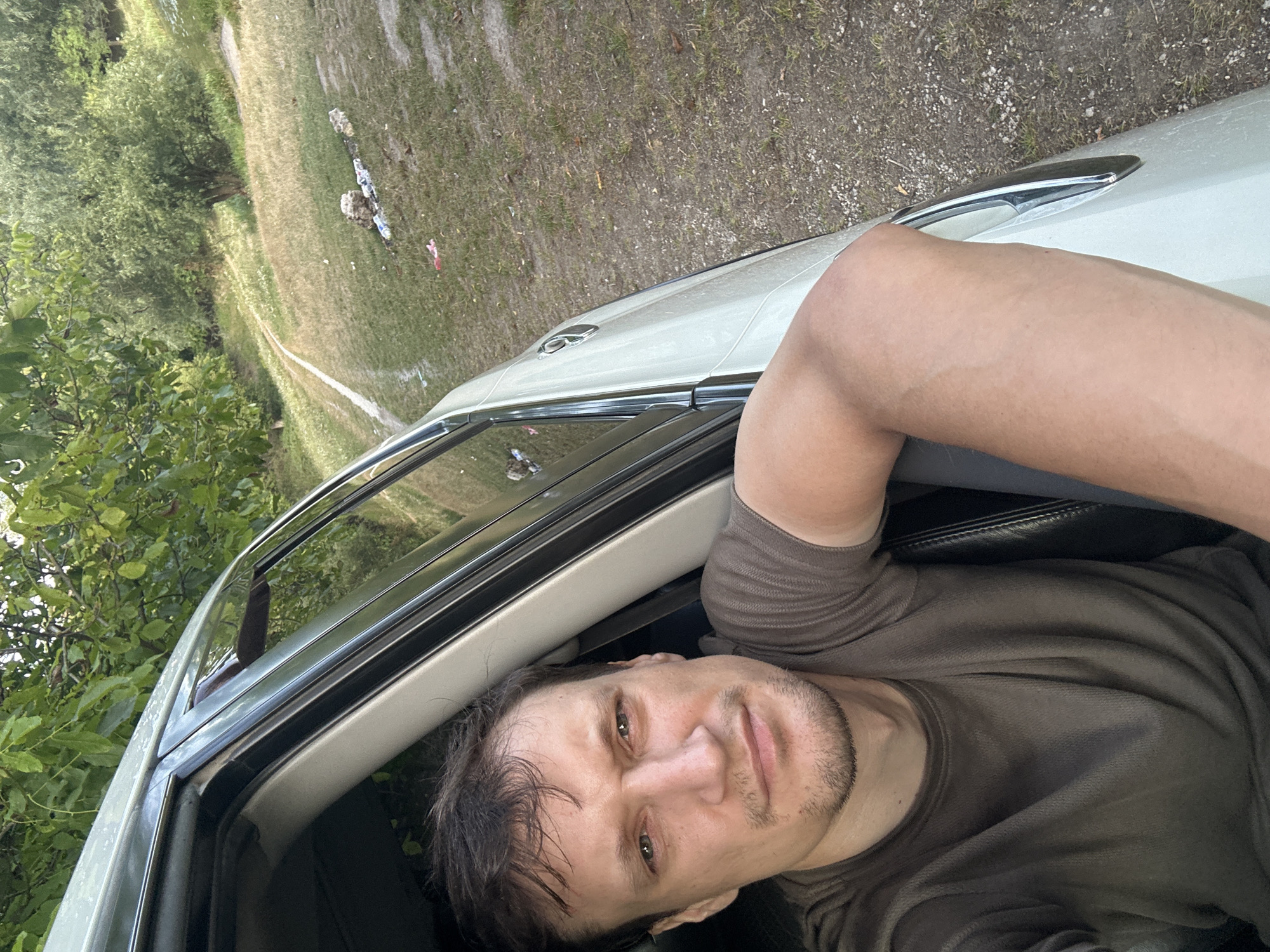 A man with short brown hair and stubble is leaning out of a car window. He is wearing a dark brown t-shirt. His right arm rests on the car door. The car is white and appears to be parked on a dirt road next to a grassy hill. The background shows trees and foliage. The scene is outdoors and appears to be in a rural area.