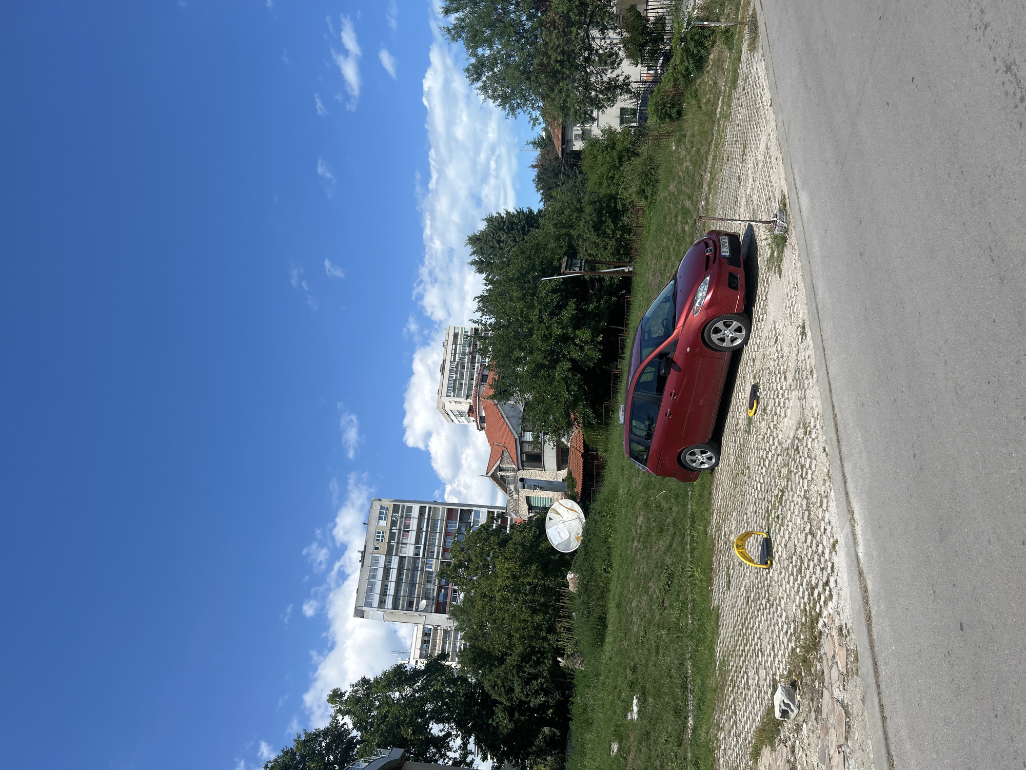 The image depicts a scene in Varna, Bulgaria, on a clear, sunny day. The sky is a vibrant blue with scattered white clouds. In the foreground, a red car is parked on a textured, light-colored paved area next to an asphalt road. The car is a compact hatchback with alloy wheels.
Behind the paved area, a strip of green grass separates it from a densely vegetated area with various trees. In the background, several buildings are visible. To the left, a tall, multi-story apartment building with numerous windows stands out. Further to the right, a smaller, older building with a red-tiled roof is nestled among the trees, and another taller building can be seen behind it. A satellite dish is positioned on the grass between the apartment building and the red-roofed structure.
Two yellow parking bollards are visible on the paved area. The overall impression is a residential or urban setting on a pleasant day. There are no people visible in the image. The time of day appears to be midday, given the bright sunlight and strong shadows. The scene is static, with no specific activity taking place beyond the parked car. A license plate on the car reads "B 696 MK".