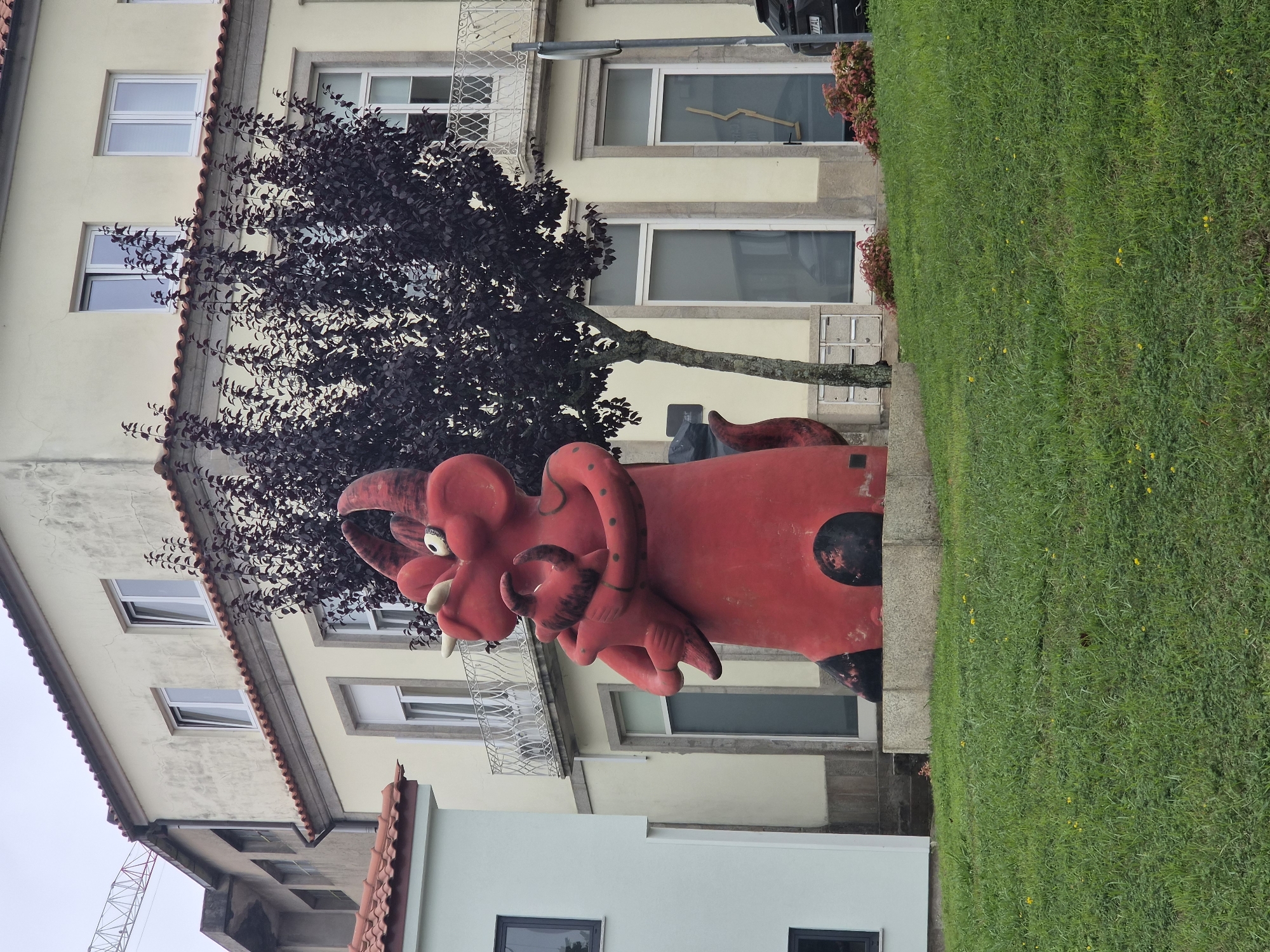 This is a ground-level, eye-level, medium shot, outdoor photograph of a large, red, concrete sculpture of a dragon-like creature embracing a smaller creature. The sculpture is situated on a grassy slope in front of a pale yellow, multi-story building with a tiled roof. The building has several windows, some with balconies. A dark-leaved tree is positioned behind the sculpture. The sky is overcast. The sculpture is the primary subject, located in the city of Barcelos, Portugal, likely in a public park or square. The sculpture is detailed, with prominent horns, eyes, and articulated limbs. The smaller creature is also rendered in red. The environment is urban, with the building suggesting a residential or commercial area. The grassy slope indicates a landscaped public space.