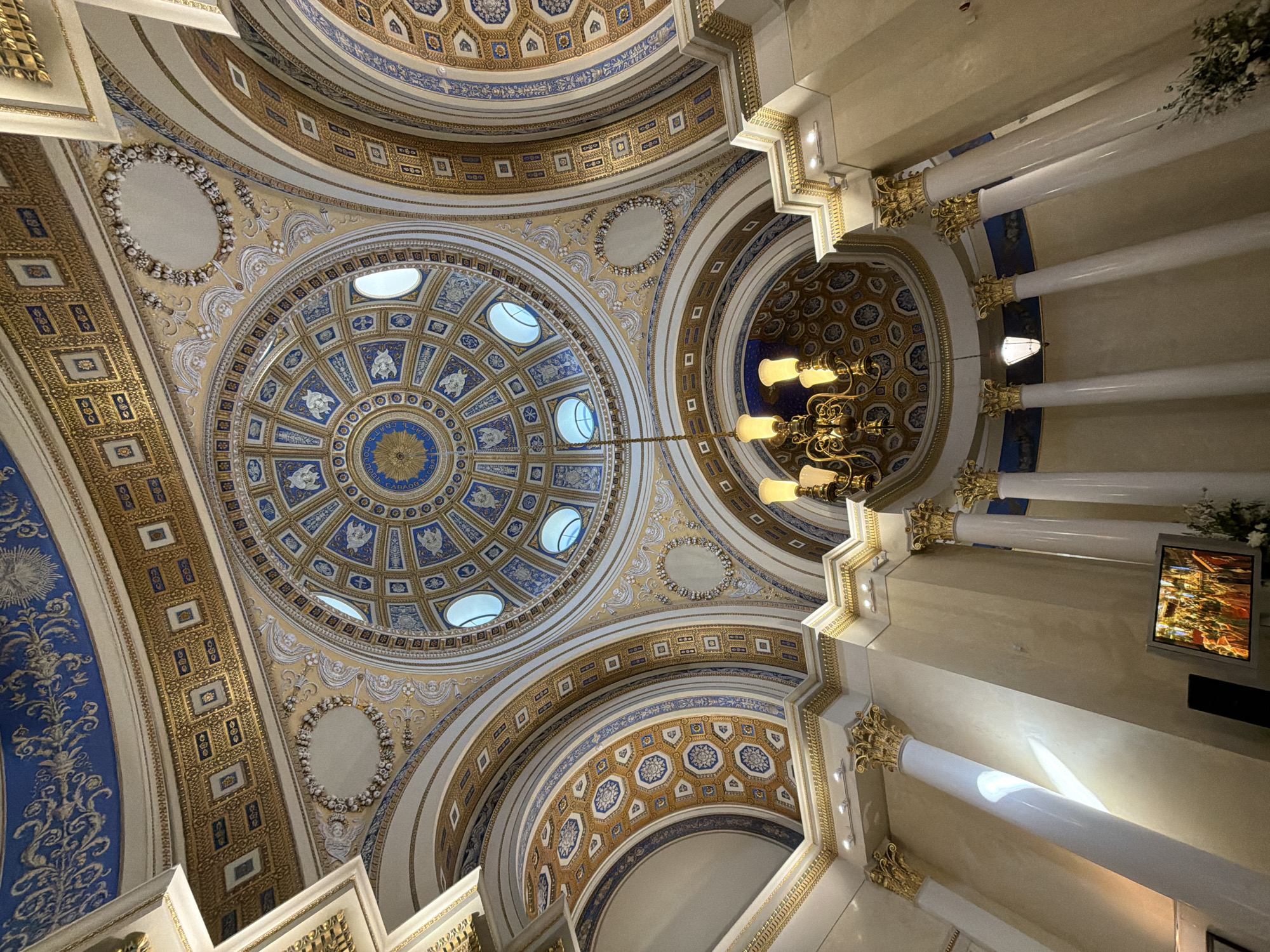 This is a low-angle, wide shot of the ornate interior of a domed building, likely a public building or place of worship. The architecture features intricate golden decorative elements on a cream-colored background, interspersed with panels of blue and gold geometric patterns and floral motifs.
Several large, white columns with golden Corinthian capitals extend upwards, supporting the domed ceiling. The ceiling itself is a complex structure with multiple smaller domes and arched sections, all adorned with detailed artwork. In the upper part of the image, a large chandelier with multiple yellow lights hangs from the ceiling.
Towards the right side of the image, a flat-screen television is mounted on the wall, displaying a colorful image, possibly of a stained-glass window or artwork. The lighting appears to be artificial, with light also entering from some small, oval windows in the ceiling. The overall impression is one of grandeur and opulence.
The location context provided is "посёлок дачного хозяйства «Архангельское», Russia," suggesting the building is located in this area. There is no visible activity or event taking place, and no discernible text in the image. The time of day is not indicated by natural light.