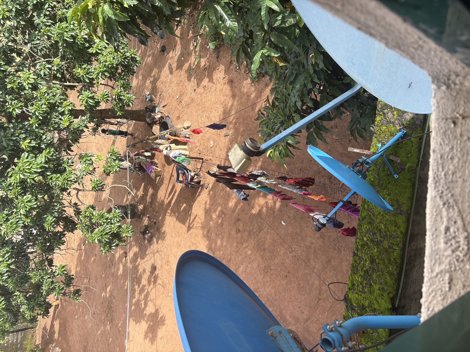 In Onitsha, Nigeria, an elevated view captures a scene with multiple elements. In the foreground, two large, bright blue satellite dishes are prominently featured, one partially obscuring the other. The dish on the left is mounted on a blue pole, with green moss growing on a concrete structure beneath it. The dish on the right is also blue and angled towards the viewer.
The background reveals a dirt ground scattered with dry leaves and shadows from trees. A clothesline is strung across the scene, laden with a variety of brightly colored clothes, including pink, red, blue, and green items. A person, appearing to be a young boy dressed in a green shirt and dark shorts, stands near a tree in the midground. Several other figures, possibly women, are gathered near a cluster of items that include chairs and buckets. A large tree with lush green leaves dominates the upper portion of the image, casting dappled shadows on the ground. Scattered on the ground are various objects such as buckets, bags, and possibly remnants of a fire pit.