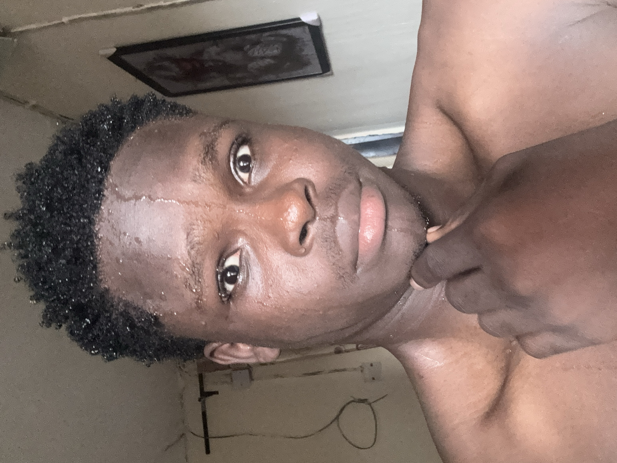 A close-up portrait of a dark-skinned man with short, curly black hair. He appears to be sweating. He is shirtless and touching his neck with his hand. In the background, there is a picture frame hanging on the wall, along with electrical outlets. The environment appears to be a room with light-colored walls. Ihiagwa, Nigeria, is not directly discernable.