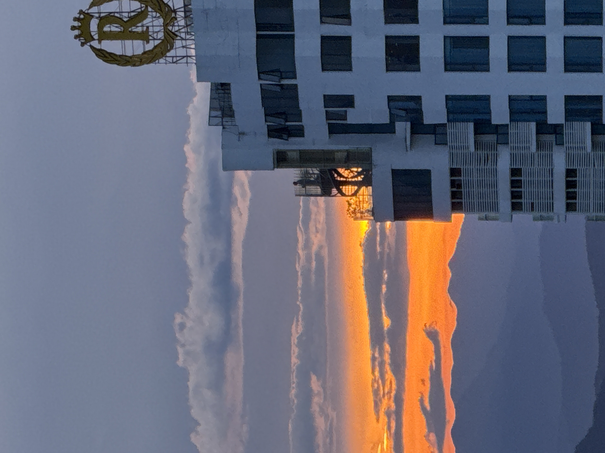 This image depicts a sunset over a city, with a prominent building in the foreground. The sky is filled with layers of clouds in shades of gray and soft purple, with a vibrant orange band of sunset light illuminating the horizon. The sun itself is a bright, diffused orange glow peeking through the clouds. In the background, a series of mountain ranges are silhouetted against the colorful sky.
The building in the foreground is a modern, white structure with a distinctive facade. It features multiple floors with rows of dark windows, some of which are open at an angle. On the upper right side of the building, a large, golden circular logo with a stylized "R" and a crown is visible. The building also has an external spiral staircase on the left side, where a solitary figure can be seen standing. The architectural style suggests it might be a hotel or a residential complex.
The overall scene is one of natural beauty juxtaposed with urban architecture, captured during the magical hour of sunset. Based on the specific visual cues in the image, such as the architectural style of the building and the mountainous landscape, this scene appears to be located in Nha Trang Ward, Vietnam. The presence of the specific "R" logo further supports this as the building is likely the Royal Beach Hotel Nha Trang.