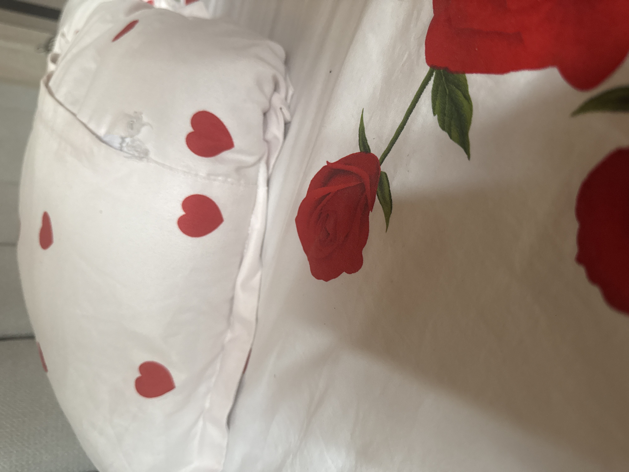 The image shows a close-up view of a white bedsheet with a repeating pattern of red hearts and red roses. The bedsheet is spread out on a bed. To the left of the image, a white pillow with red hearts is visible, partially covered by the bedsheet. A faint seam runs vertically along the left side of the pillow. The lighting suggests an indoor setting, possibly a bedroom. The focus is on the fabric's pattern and texture, with soft shadows indicating the folds of the material. There are no people or any discernible activity taking place. The scene is static and appears to be a detail shot of bedding. There is no visible text in the image. The context provided of Tashkent, Uzbekistan, does not offer any specific visual cues within the image itself.