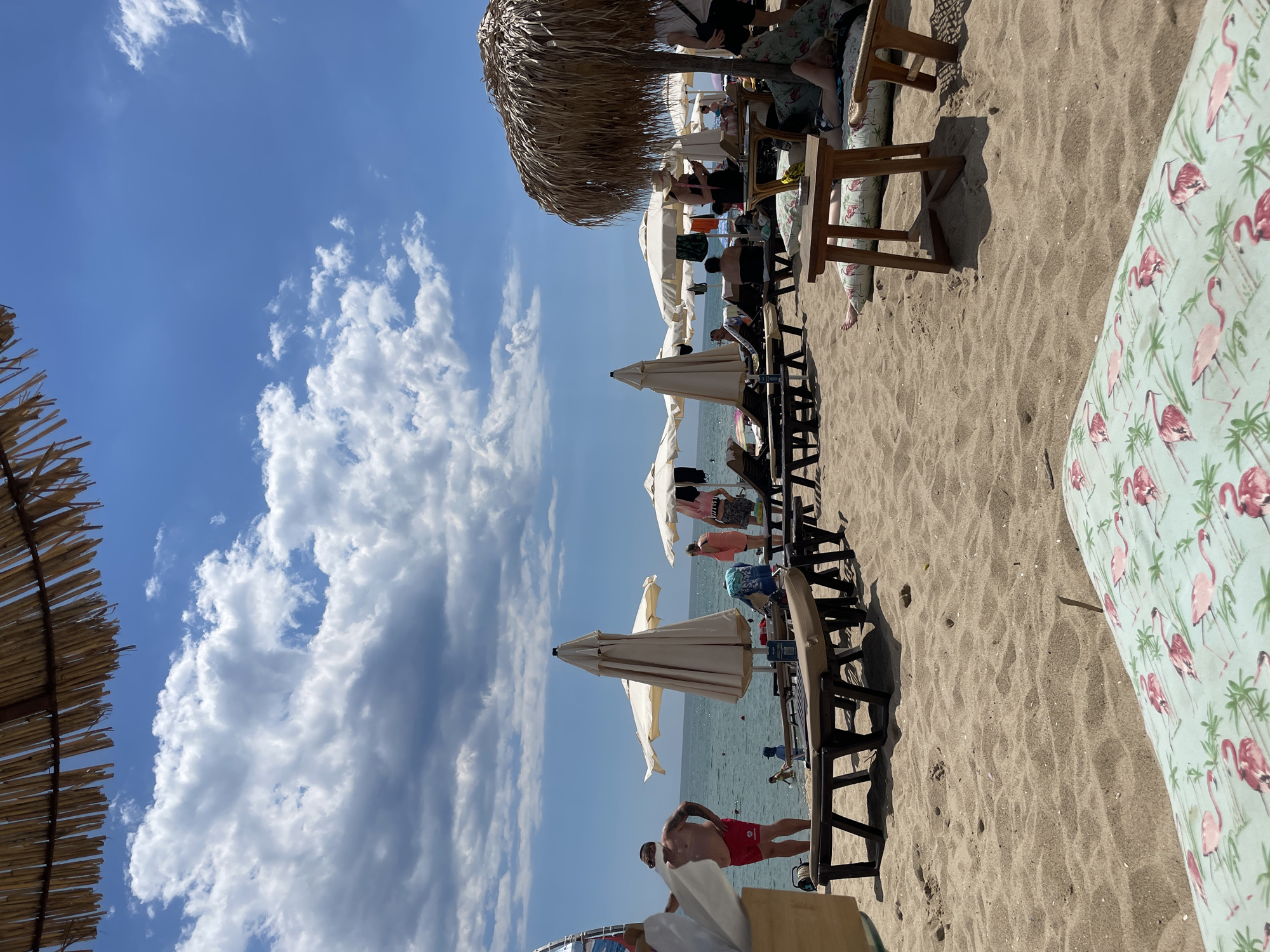The image depicts a crowded beach scene on a sunny day in Obzor, Bulgaria. The foreground is occupied by a sandy beach with several sun loungers and cushions with a flamingo pattern. In the mid-ground, numerous beach umbrellas and sun loungers are set up. People are scattered across the beach, some relaxing on loungers, others walking or standing near the water's edge. The sea is visible in the background with calm waters and a few people wading in it. The sky is partly cloudy with a bright blue expanse. A man in red shorts stands near the water, facing away from the camera. Several beach umbrellas, some open and some closed, are positioned strategically on the sand. A straw-thatched umbrella is prominent on the right side of the image. The overall atmosphere suggests a typical summer day at a popular seaside resort.