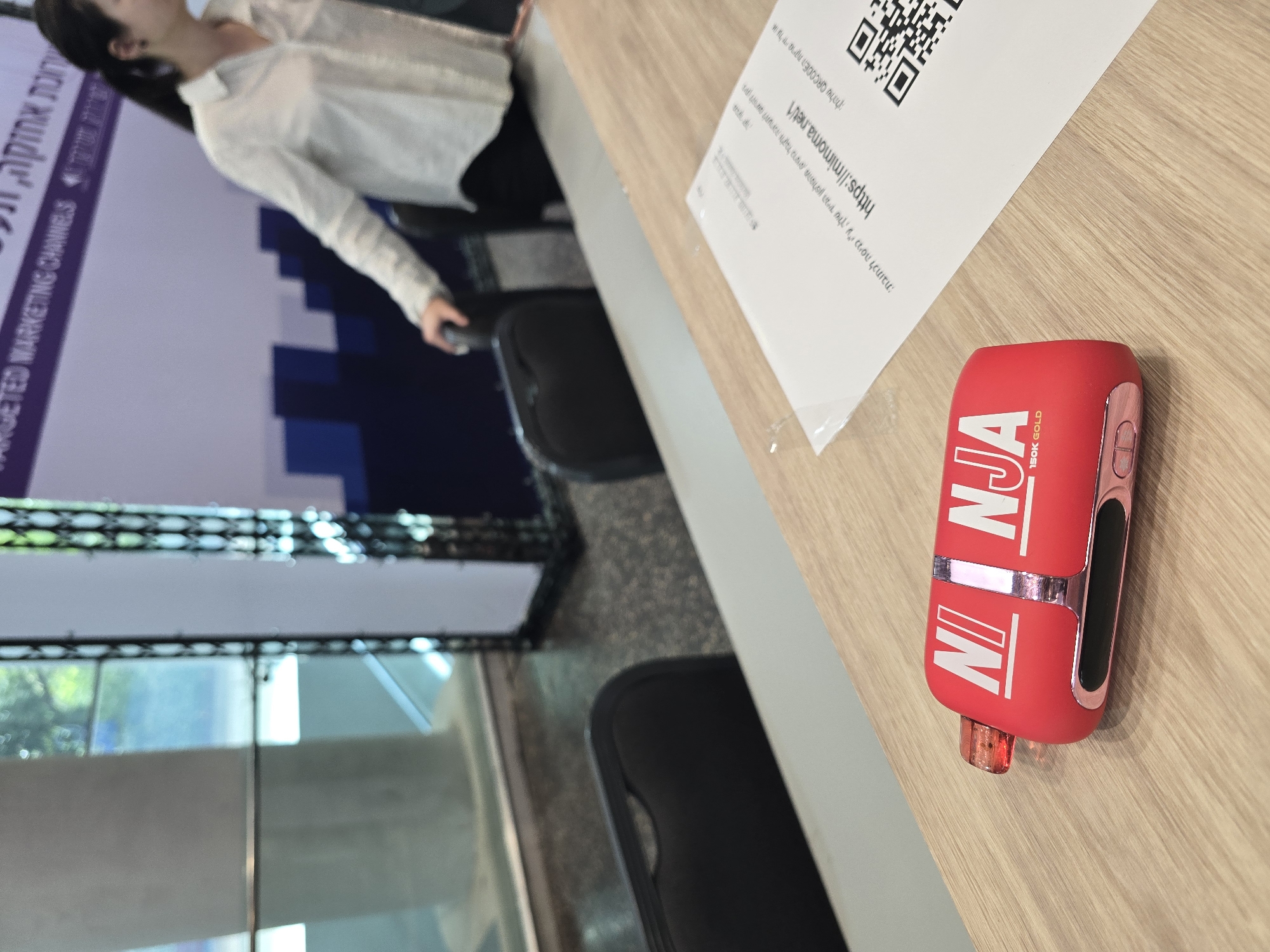 The image shows a red "Ninja 150K Gold" vape pen on a wooden table next to a piece of paper with a QR code and a website address ("https://mimoma.net/1") written in Hebrew. The paper also has Hebrew text discussing entering the address to receive gifts from the device. In the background, a woman in a white shirt is seated. A banner, also with Hebrew writing ("TARGETED MARKETING CHANNELS"), is visible, along with a glimpse of the surrounding environment, including what appears to be windows or a glass facade. Based on the Hebrew text and general appearance, the location is likely in Tel-Aviv, Israel.
