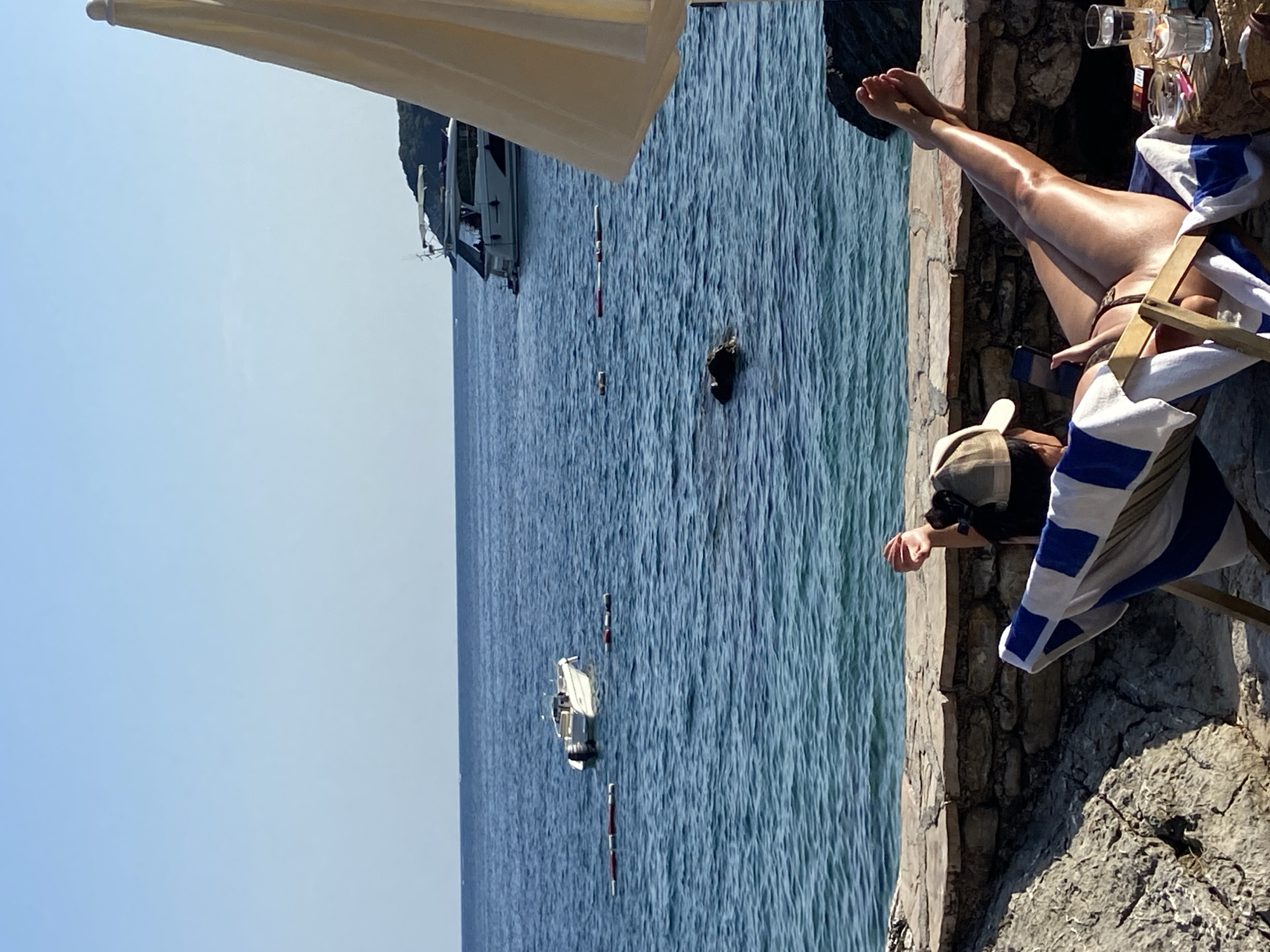 The image depicts a person relaxing on a lounge chair on a rocky shoreline overlooking the sea. The person is wearing a bikini and a baseball cap, and they are holding a phone. The sea is a deep blue and calm, with a few boats in the distance. A large umbrella provides shade. The shoreline is made of rough stones and a low stone wall. The scene appears to be a beach or a waterfront area in Prijevor, Montenegro, suggesting a tranquil vacation setting.