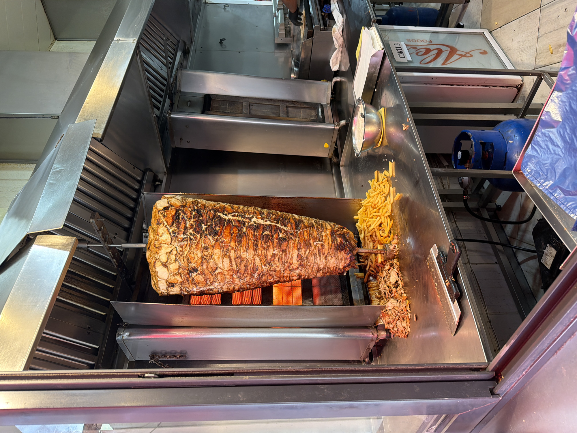 The image shows a shawarma or gyros grill inside a street food establishment. A large cone of meat is mounted on a vertical rotisserie and is being cooked by infrared heaters. The meat is carved and accumulating on the counter next to some French fries. The establishment appears to be located in Plovdiv, Bulgaria, based on the "Alex Foods" sign, CКЛ13 on the sign next to the food. A blue gas tank is located nearby.
