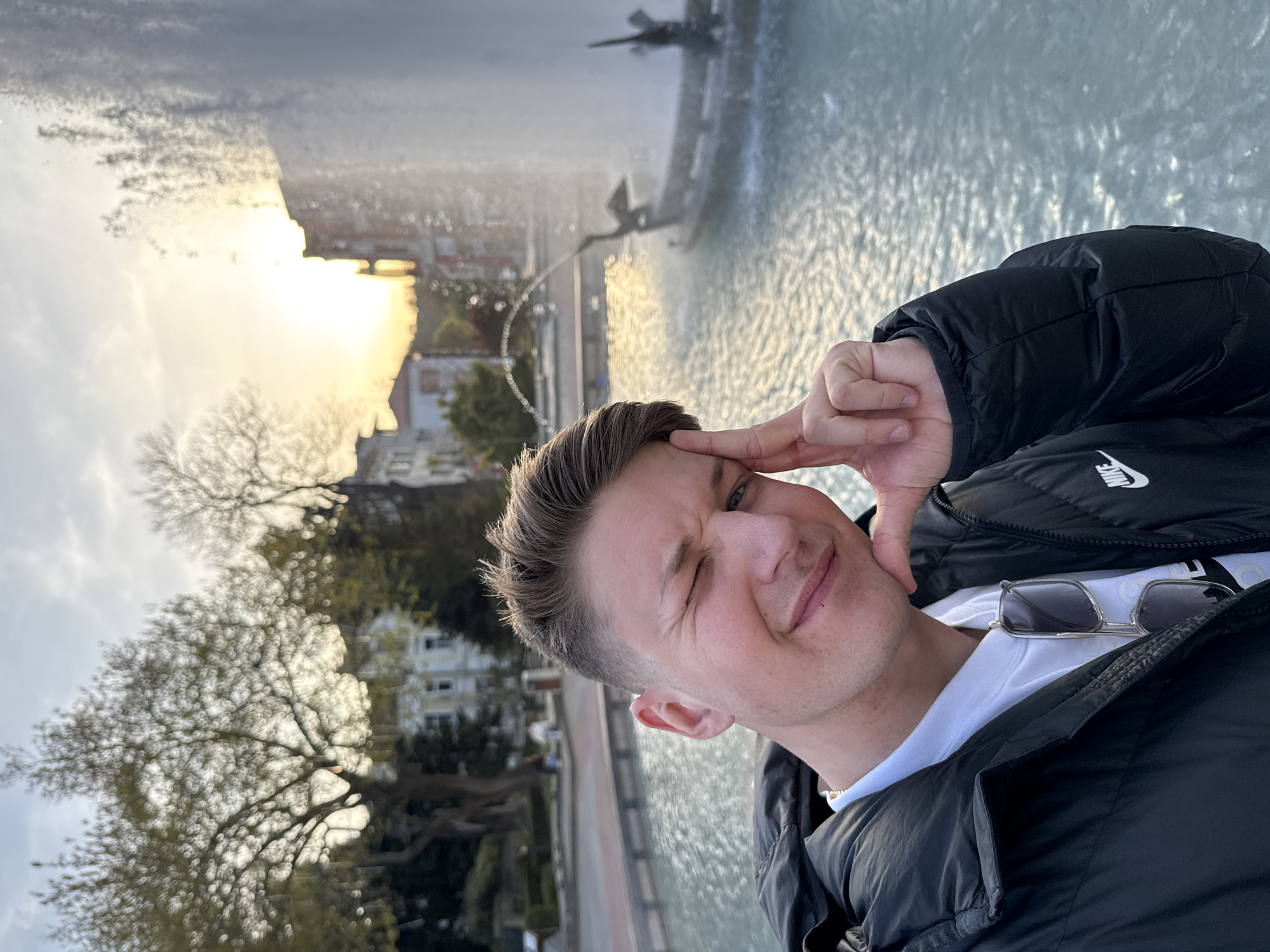 A young man poses for a selfie in Plovdiv, Bulgaria, at sunset. He is in the foreground, winking, with his finger on his forehead, wearing a black Nike jacket over a white t-shirt, with sunglasses resting on his chest. Behind him is a large, ornate fountain, with the setting sun in the background, illuminating buildings and trees. The sky is partly cloudy.