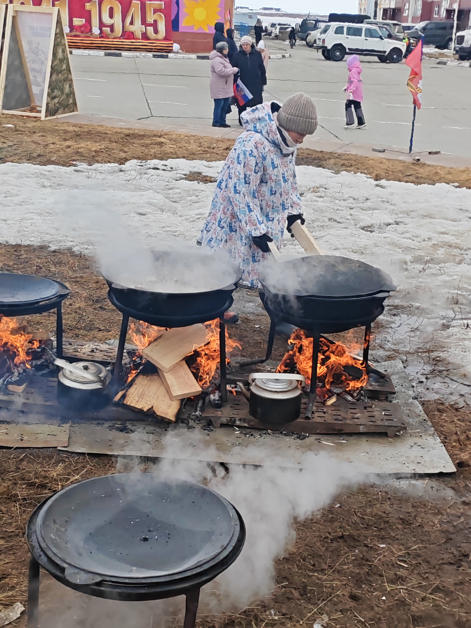 The image, taken in Ugolnye Kopi, Russia, shows a person stirring a large pot over an open fire. The person is wearing a patterned coat and a knitted hat. There are two other similar pots on stands nearby, one of which is covered. Fires are burning beneath each pot, with kettles placed close to the flames. Steam rises from the active cooking pots. The ground is covered with patches of snow and dry grass. In the background, there is a banner with the year "1945," along with other people, vehicles, and buildings, suggesting a public event.
