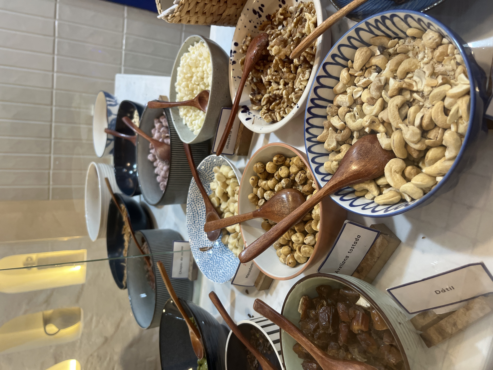 The image shows a selection of toppings or ingredients, possibly for a dessert bar or yogurt topping station, presented in a buffet-style arrangement in Puebla, Mexico. There are several bowls containing different items such as white chocolate chips, nuts (walnuts, cashews, hazelnuts, macadamia nuts), and dates. Each bowl has a small wooden spoon for serving. Small labels identify the contents of each bowl, including one labeled "Dátil" (date in Spanish) and "Avellana tostada" (toasted hazelnut). The bowls are arranged on a white countertop, and there's a glass partition to the left, possibly part of a display case. The angle suggests the photo was taken from a slightly elevated position, looking down at the arrangement. The background includes neutral-colored tiled walls and glimpses of other containers or display elements.