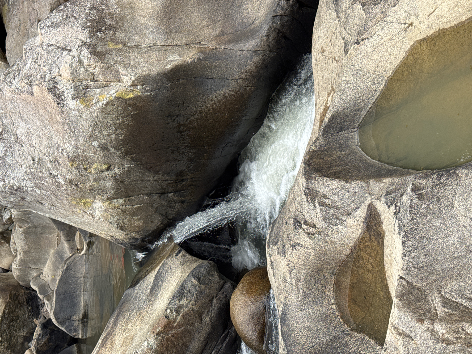 This is an outdoor, eye-level, close-up photograph of a narrow waterfall cascading between large, rough, weathered rocks. The water is white and frothy as it tumbles down, creating a dynamic contrast with the stillness of the stones. The rocks themselves are of varying sizes and shapes, with some featuring smooth, polished surfaces and others appearing more jagged and eroded. There are also small pools of still water collected in natural depressions on the rocks in the foreground, reflecting the muted light. The overall scene depicts a natural water feature in a rocky environment. This location is likely within the natural landscape of Ninh Hoa City, Vietnam, though specific landmarks are not identifiable. There are no people or animals visible in the image.