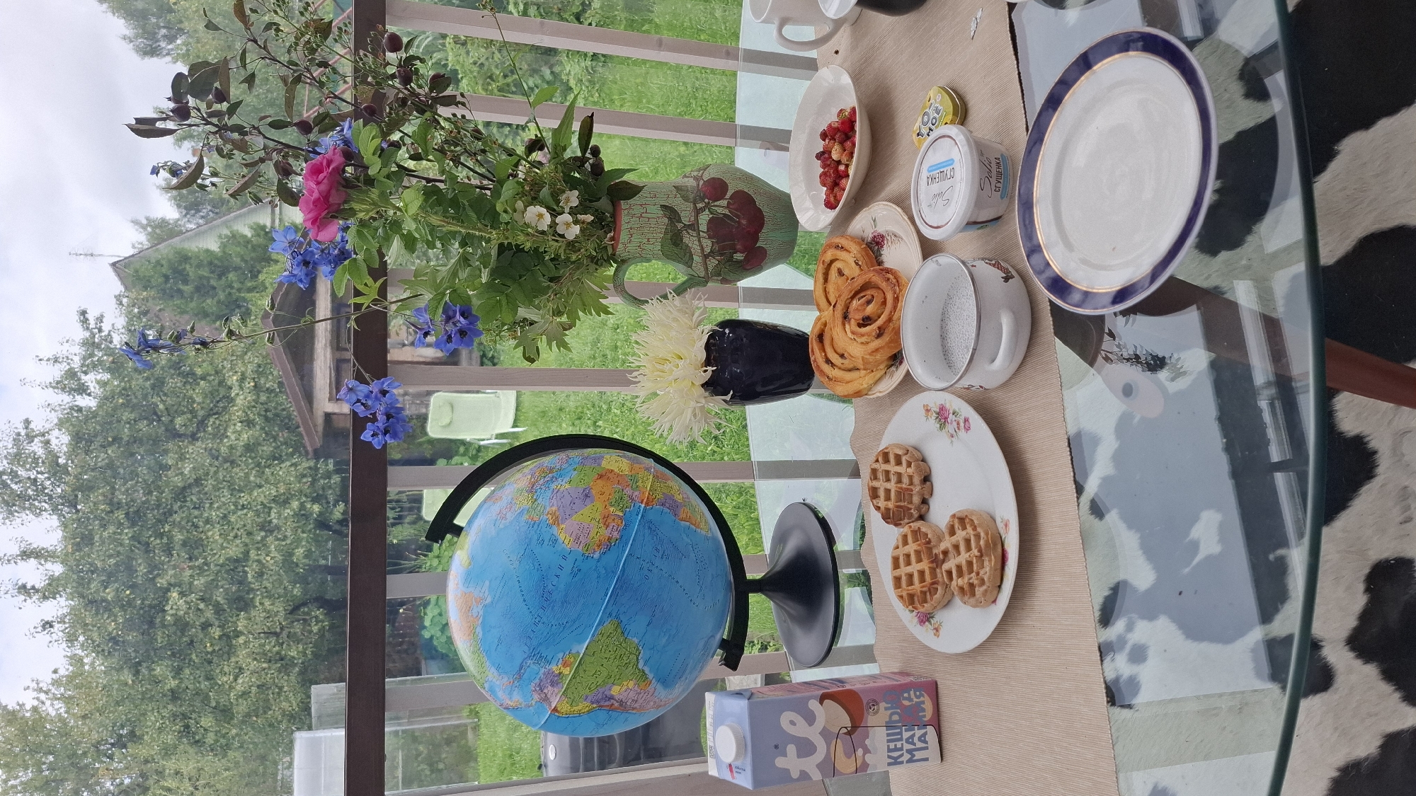 On a glass-topped table on a balcony, a breakfast spread is laid out with a globe in the background. The table is covered with a light-colored placemat. To the left, a carton of milk with Russian text and an illustration of a cat is placed next to a blue and green globe. In the center, a plate holds three waffles, a small white bowl contains a chia seed pudding, and another plate has two cinnamon rolls. To the right, a small white bowl is filled with wild strawberries, and a tub of condensed milk sits nearby. A bouquet of flowers, including pink roses and blue delphiniums, is arranged in a green pitcher decorated with cherries. The background reveals a backyard scene with trees, a wooden shed, and a house, indicating the location is likely in a suburban or rural area of Орбита Снт (терпигорьево), Russia, during daytime with an overcast sky.