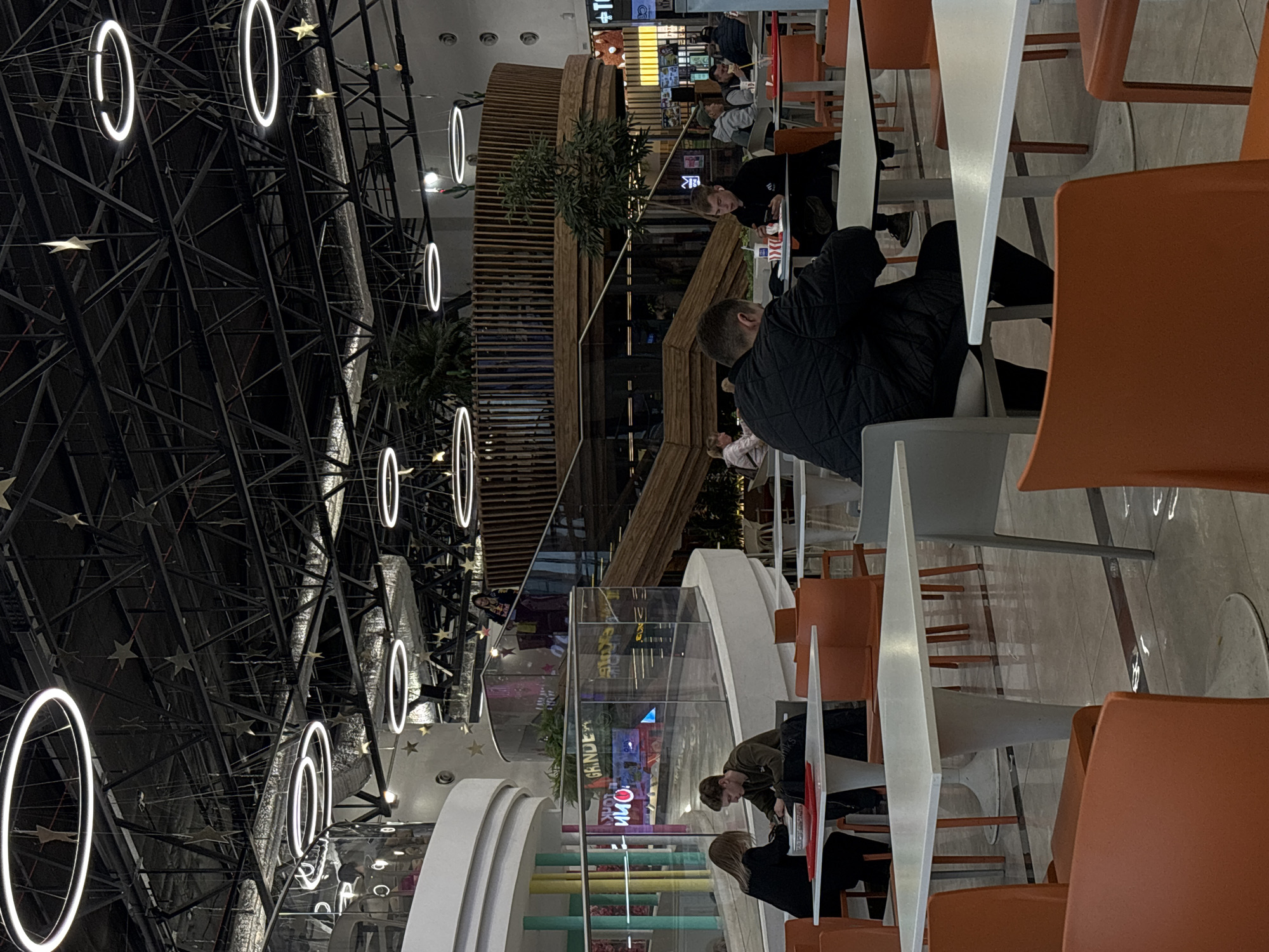 This photograph, taken in Perm, Russia, showcases an indoor scene, possibly within a shopping mall or food court. The area features high ceilings with complex metal framework adorned with circular LED lights. Several individuals are seen seated at tables, some appearing to be eating or using electronic devices. The furniture consists of white, triangular-shaped tables and orange chairs. A multi-level structure made of light brown wood is also visible, with indoor plants on its upper levels. Signage in both English and what appears to be Russian script suggests a diverse environment.