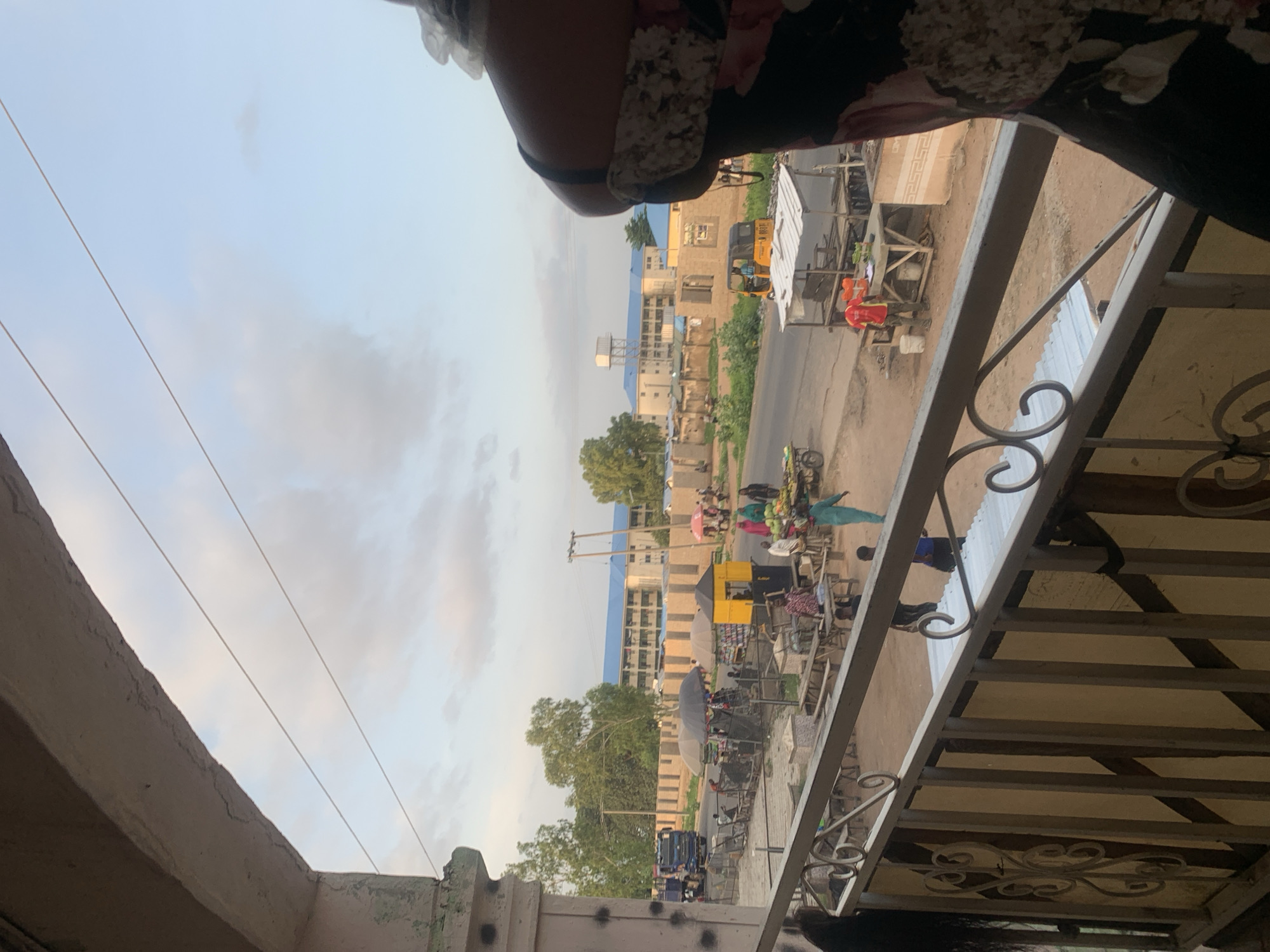 This image depicts a street scene in Jalingo, Nigeria, viewed from an elevated vantage point, likely a balcony. In the foreground, a section of a railing with decorative metalwork is visible, with part of a person's torso and shoulder on the right side, wearing a patterned garment.
The middle ground shows a bustling street with market stalls and people engaged in various activities. Several umbrellas provide shade over the stalls, where goods are displayed. A yellow kiosk with a black roof is prominently situated. People are seen walking, interacting, and some appear to be vendors. A fruit cart laden with watermelons and other produce is visible on the right side of the street. A yellow three-wheeled vehicle, possibly an auto-rickshaw, is parked on the road. Further down the street, a blue truck is parked.
In the background, buildings with blue roofs suggest an urban environment. Trees line the street and are interspersed among the structures. The sky above is a pale blue with scattered clouds, indicating it is likely late afternoon or early evening. The overall scene suggests a vibrant marketplace and a typical day in a Nigerian city.