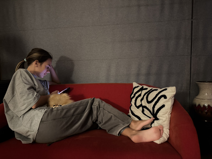 The scene depicts an indoor setting in Warsaw, Poland. A female individual, with light brown hair tied in a ponytail, is seated barefoot on a vibrant red upholstered sofa, dressed in a grey t-shirt and grey sweatpants. She is focused on a dark-colored smartphone held in her hands, its screen emitting light onto her face. A small, fluffy, light brown dog, consistent with a Pomeranian-type breed, rests on her lap. A cream-colored decorative cushion featuring a black abstract linear pattern is positioned on the right side of the sofa, behind her feet. The background wall is dark grey with visible horizontal paneling or texture. On the far right, a decorative vase or urn with a textured, light-colored body and a dark red or maroon patterned lower section is partially visible. The overall lighting of the environment is dim.