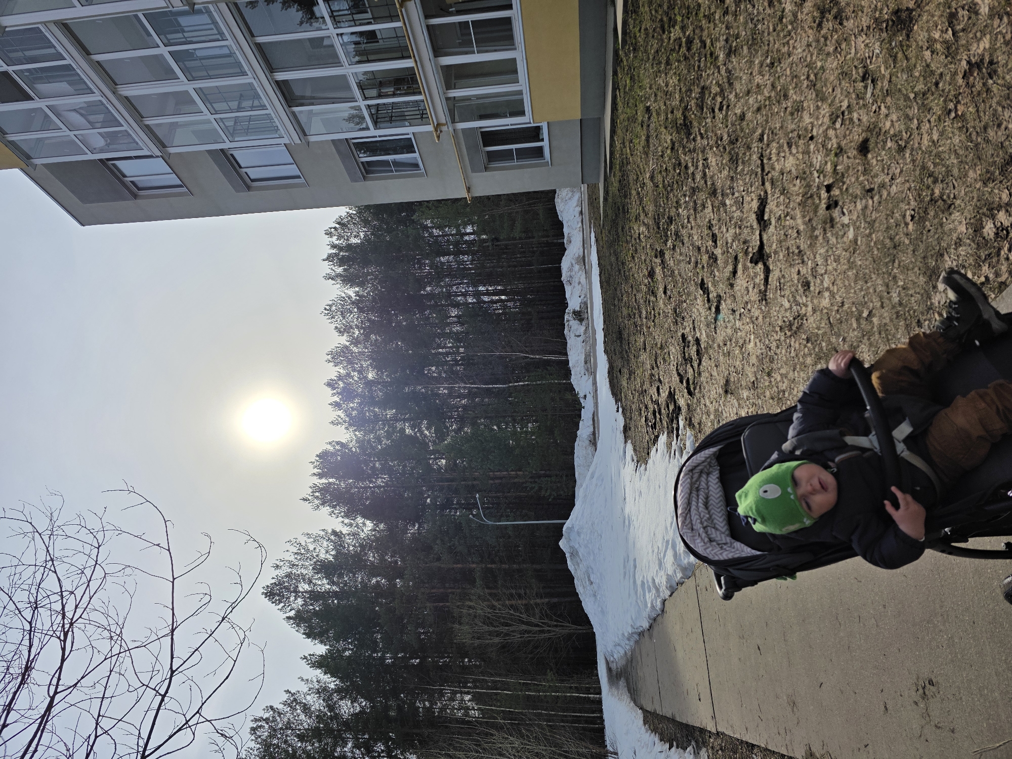 A child wearing a green hat is in a stroller on a sidewalk. To the left of the sidewalk is a bank of snow and beyond that is a wooded area. Further to the left, there is a tall building. The sky is visible with the sun shining through the haze. The location is the city of Chaikovsky, Russia.