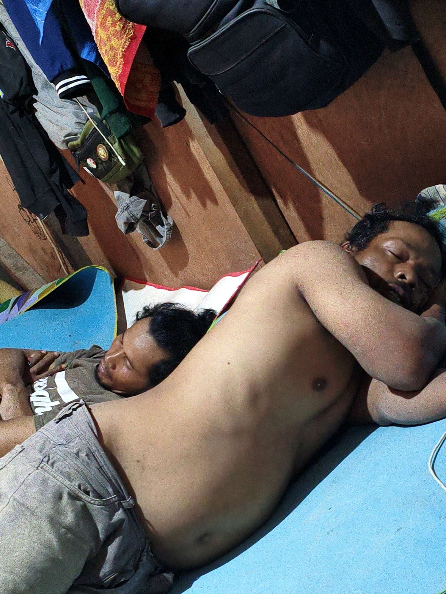 The image shows two men sleeping next to each other on a blue mat. The man in the foreground is shirtless and wearing grey pants, while the man behind him is wearing a brown shirt with white stripes. The background is a wooden wall with various items hanging on it, including clothing, a bag, and a colorful blanket. The lighting suggests it might be indoors and either daytime or nighttime, as there are shadows cast on the wall. The overall mood appears to be relaxed and peaceful, with the men in a deep sleep. There is some faint text visible on the shirt of the man in the background, which reads "yba". The setting appears to be a simple living space, possibly in a home or dormitory. The presence of the mat and the clothing suggest a casual and informal environment.