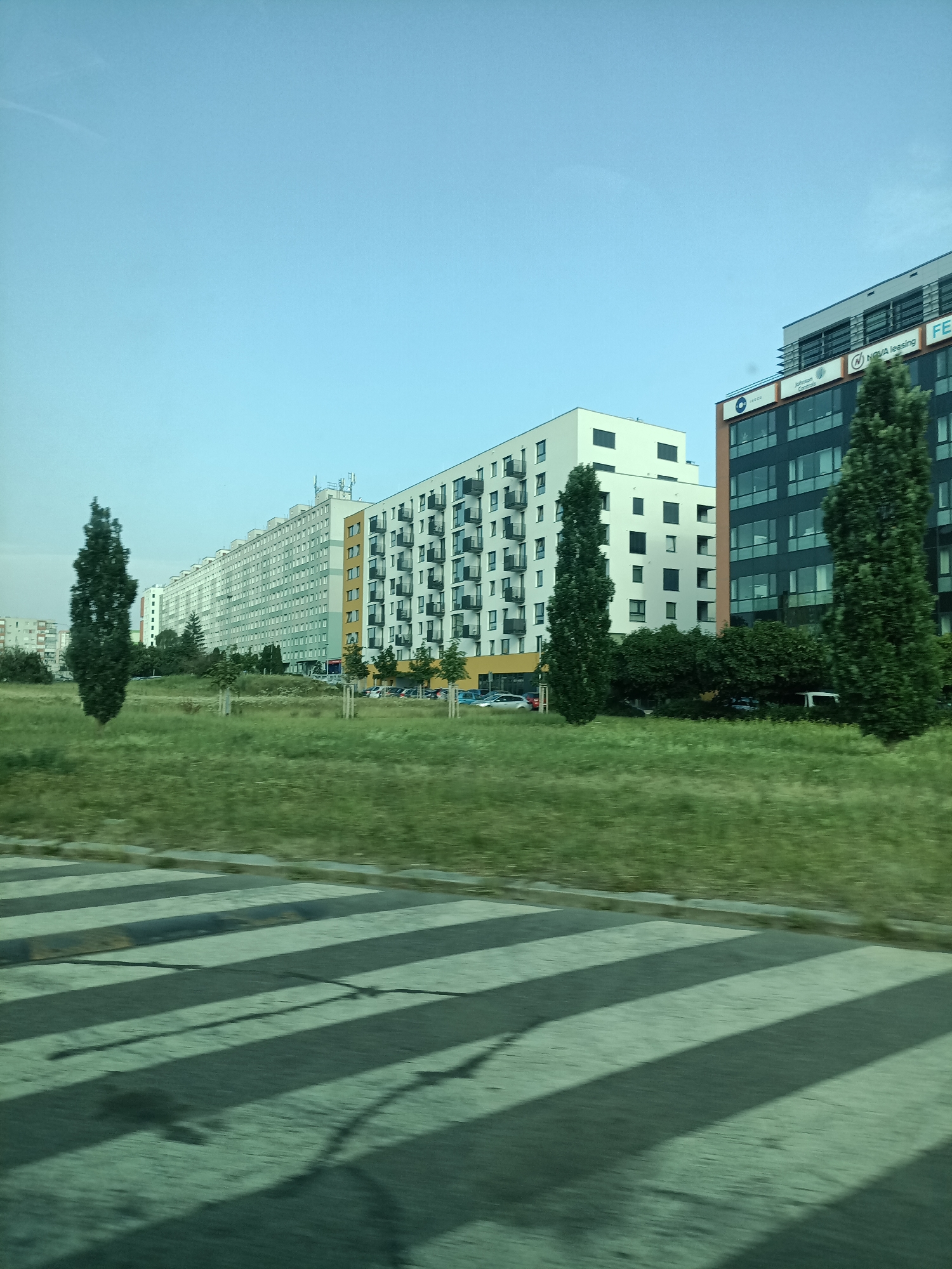 This media file depicts an outdoor scene in Prague, Czechia, under a clear, pale blue sky suggesting daytime. The foreground features a blurry crosswalk with alternating dark and light stripes, indicating a street or road. Beyond the crosswalk, a grassy area is visible, with several young trees planted in a row.
In the mid-ground and background, a series of modern buildings dominate the landscape. To the left, a long, light-colored apartment building stretches across the frame, its facade punctuated by numerous windows and balconies. To its right, a more contemporary white building with dark-framed windows stands out. Further to the right, a darker, multi-story building with a glass facade and orange accents is prominently featured. This building displays visible text, including a blue logo with white lettering that appears to read "N NOVA leasing" and "FE". Another smaller logo is also visible above this text.
The overall scene suggests an urban environment with a mix of residential and commercial architecture. The presence of trees and grass indicates a planned urban development, possibly a residential area or a business park. The blur in the foreground suggests the image was taken from a moving vehicle, capturing a fleeting glimpse of the cityscape. There are no people or overt activities depicted, focusing instead on the architectural elements and the urban setting. The weather appears to be pleasant, given the clear sky.
