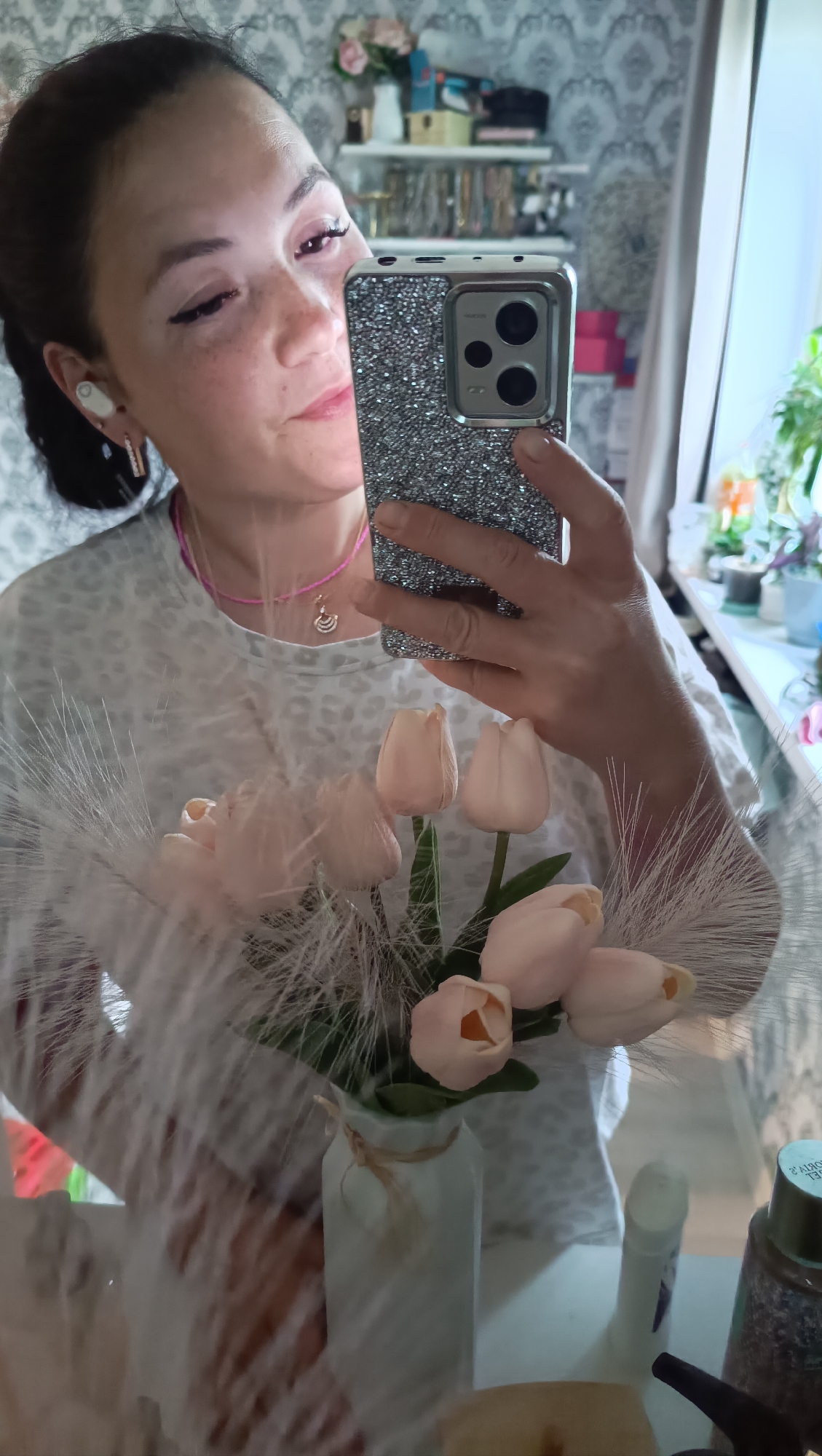The image depicts a woman holding a smartphone in front of a mirror, taking a selfie. She is wearing a white, leopard-print shirt and a pink beaded necklace with a small pendant. She has on white wireless earbuds and appears to have false eyelashes. In her other hand, she is holding a bouquet of light pink tulips and some fluffy white plumes in a vase. The background shows shelves with various items, including what appear to be decorative objects and possibly makeup. The overall setting suggests a home environment, likely a bedroom or dressing area. The image does not provide any specific geographical clues to pinpoint its location within Chaikovsky, Russia, or any other city.