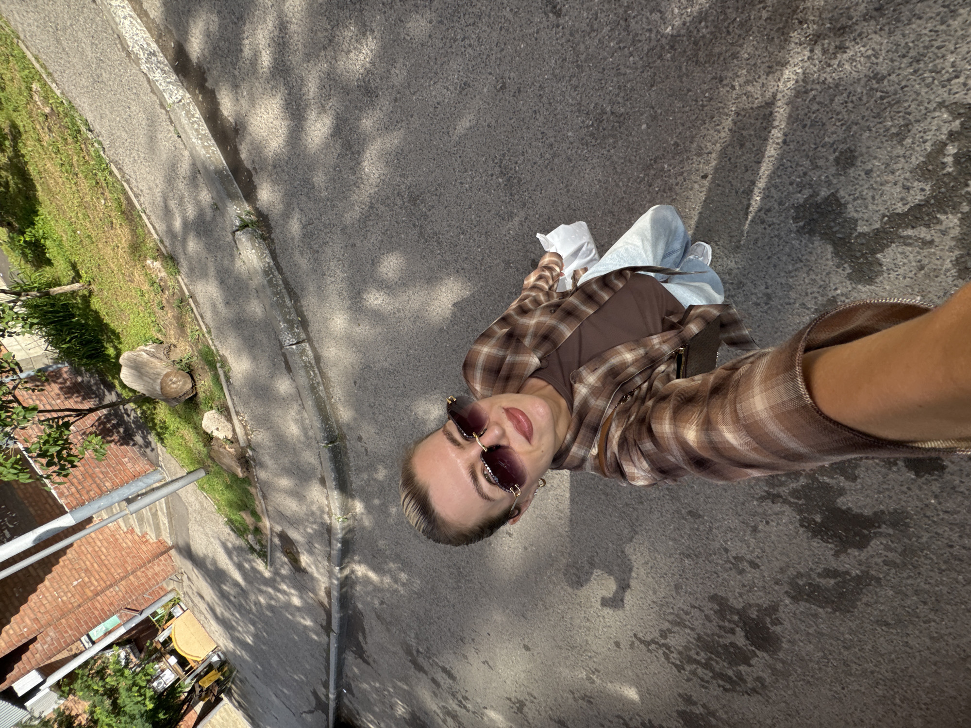 A woman is captured in a selfie taken outdoors on a sunny day in an urban environment. She is standing on an asphalt surface, likely a street or walkway, with dappled sunlight and shadows cast from overhead foliage.
The woman is the central subject of the image. She is wearing a brown plaid shirt over a brown t-shirt, light blue jeans, and white sneakers. She has on large, fashionable sunglasses with a rose gold frame. Her hair is pulled back. She is holding a white plastic bag in her right hand, and her left arm is extended to take the photo. Her expression is a slight smile.
The background reveals elements of an urban setting. To the left, a brick building is visible, with some greenery and what appears to be a construction or scaffolding structure. Further to the left, a yellow object, possibly a bike or vehicle, is partially visible. A concrete curb runs along the edge of the asphalt. In the mid-ground, there is a grassy area with a fallen log and more vegetation. The overall impression is a casual outdoor scene in a city.