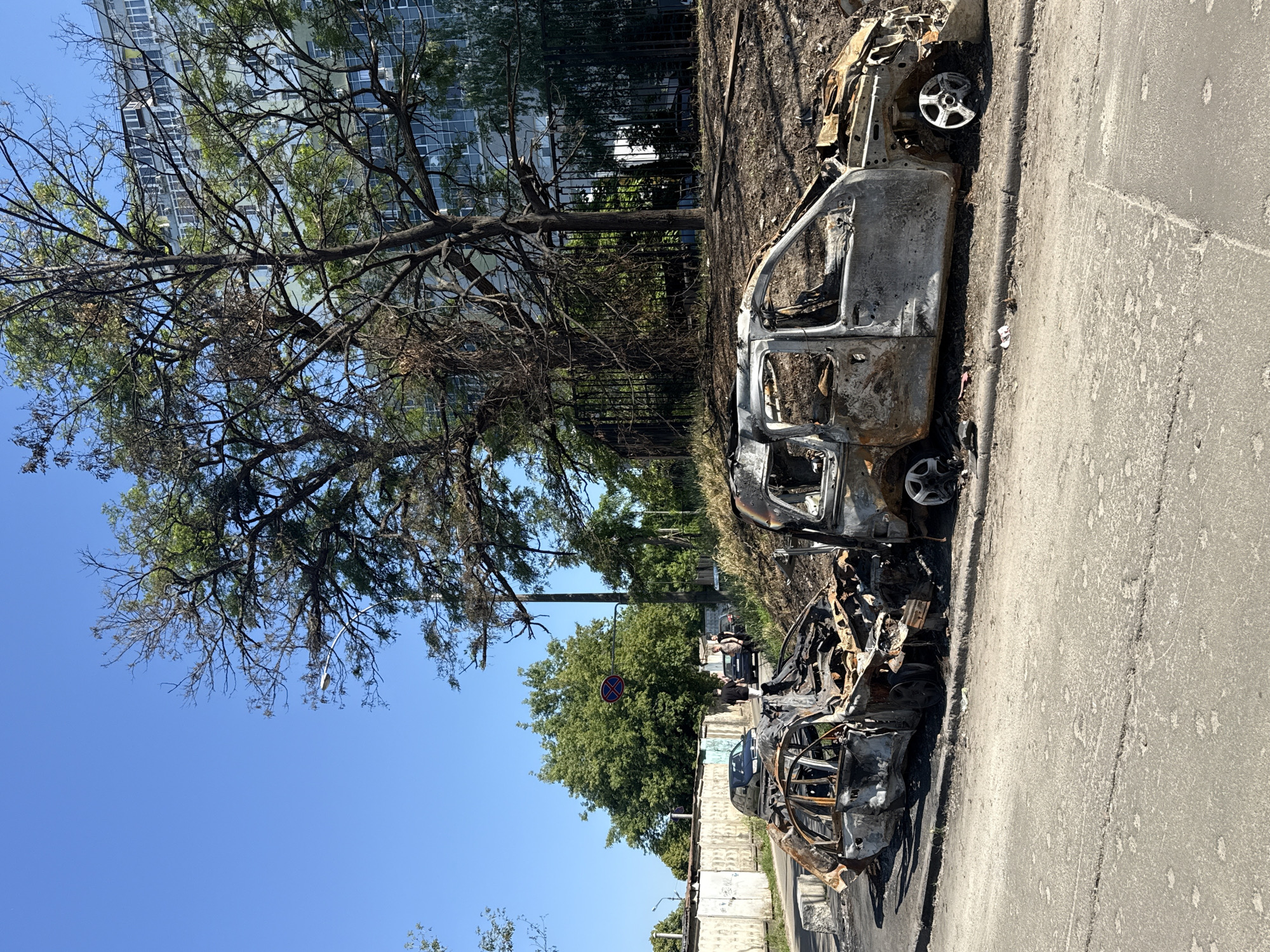 In Kyiv, Ukraine, two severely burned-out cars are parked on the side of a road. The car in the foreground, a light-colored SUV, has lost its roof and most of its interior, with exposed metal framing and melted plastic. Its tires are partially intact, and the wheels are visible. To its left, a smaller, darker-colored car is also extensively damaged, appearing to be fused to the ground in some areas, with its entire structure warped and blackened. The road surface is asphalt, marked with white lines, and a concrete curb separates the road from the embankment. Behind the cars, a grassy slope rises, leading to a dense line of trees. In the background, a modern, multi-story building with numerous windows stands behind a black metal fence and more trees. Several individuals are visible in the distance, standing near other vehicles on the road, some appearing to observe the scene. The sky is clear and blue, suggesting a sunny day.