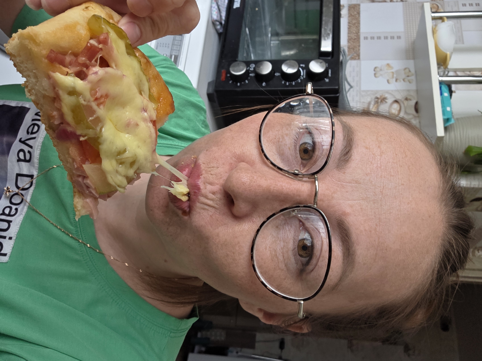The image depicts a close-up, eye-level view of a woman actively eating a piece of food within what appears to be a residential kitchen in Chaikovsky, Russia. The woman, with brown hair styled back and visible freckles on her forehead, wears round, tortoiseshell-patterned eyeglasses and a plain green t-shirt with a delicate gold-colored necklace. Her brown eyes are directed towards the camera. She is shown with her mouth partially open, taking a bite from a small, golden-crusted baked item resembling a personal pizza or calzone, which she holds with her left hand. A prominent string of melted yellow cheese stretches from her mouth to the food, which is topped with diced red tomatoes, green pickle slices, and sliced meat. The background reveals a kitchen environment, with a black countertop oven featuring silver control knobs visible to the left. Above the oven, a white shelf holds a yellow-footed white cup and a small blue package. The wall behind these elements is tiled with a light-colored pattern featuring coffee-related motifs. Part of a white appliance with a control panel is visible in the lower left background.