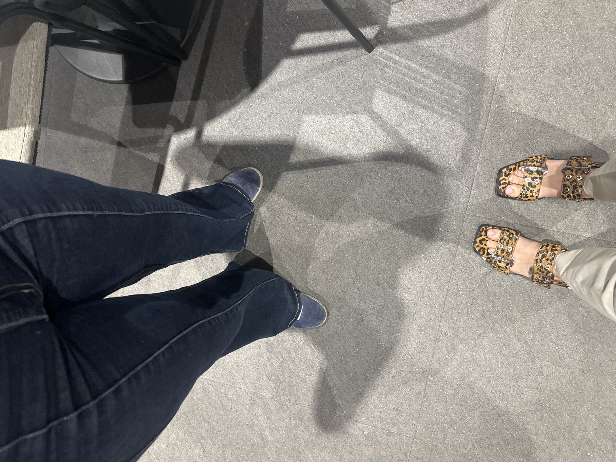 The image shows the lower halves of two people standing on a gray tiled floor. One person is wearing dark blue denim jeans and blue suede shoes. The other person is wearing khaki pants and leopard print sandals with metal buckles. A chair is visible in the upper left corner of the frame. The location is likely indoors on a smooth, light-colored floor. Given that there is no information about the location in the city of Huixquilucan de Degollado, Mexico, and it can't be determined from the image, that information will be omitted.