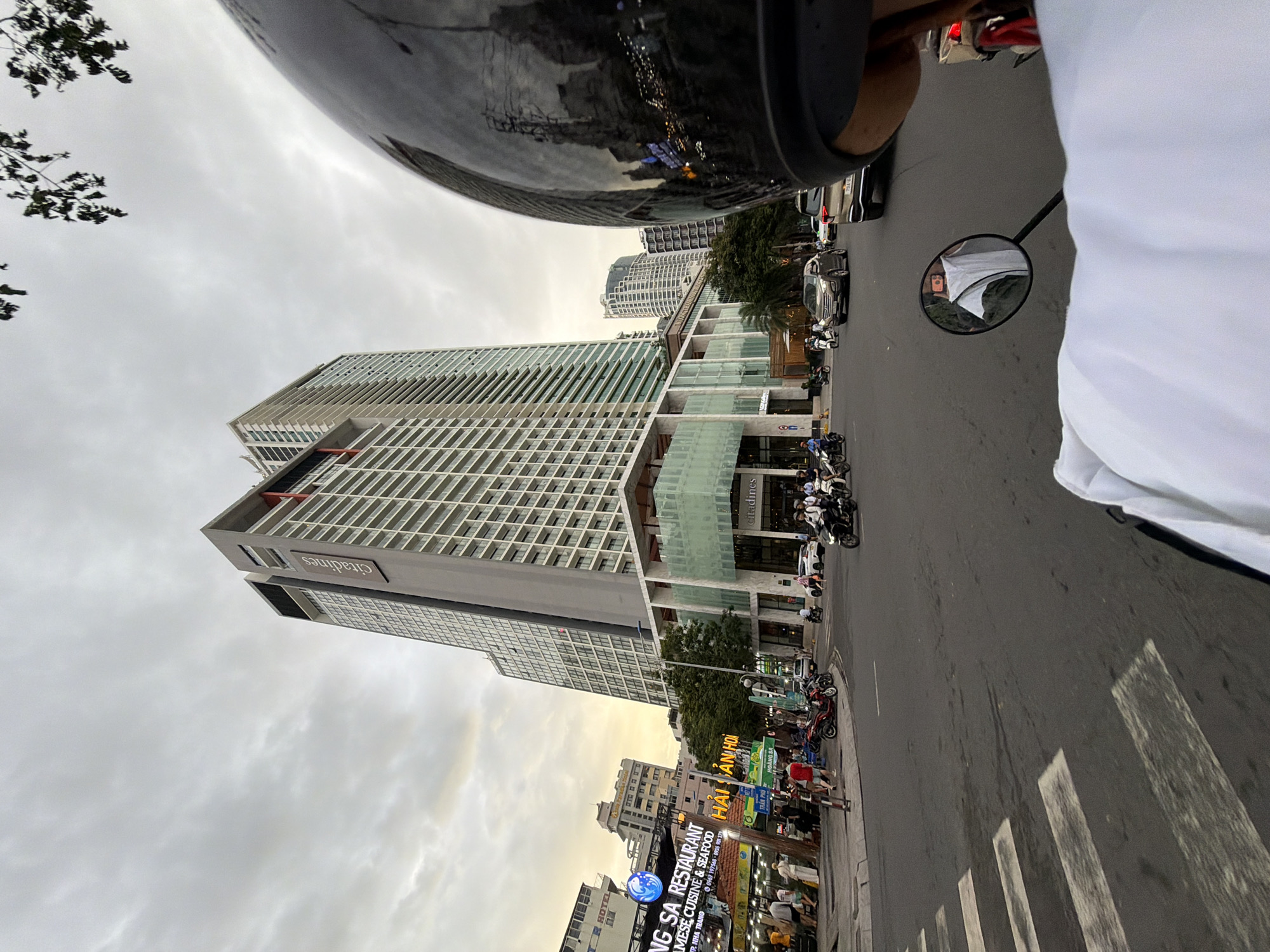 The image captures a street scene in Nha Trang, Vietnam, with a prominent, modern skyscraper dominating the background. The skyscraper, branded with "Citadines," features a glass facade and appears to be a hotel or apartment complex. In the foreground, a person on a motorcycle is looking forward, with a partial view of their helmet and a white garment they are wearing. The street is busy with traffic, including several other motorcycles and a few cars. Pedestrians are visible on the sidewalk, some near restaurants and shops. The sky is overcast, suggesting either early morning or late afternoon. The overall impression is a snapshot of urban life in a coastal Vietnamese city.