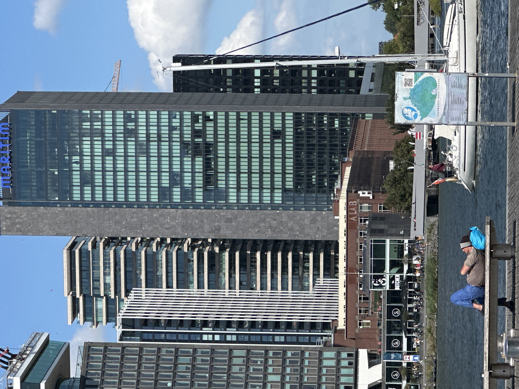 In Amsterdam, Netherlands, a man rests on a bench in the foreground, his head supported by a blue pillow, eyes closed. Behind him, a body of water is visible, with a sailboat docked. Further in the background, a row of modern buildings dominates the skyline. The tallest building, a glass skyscraper, has "MARITIM" written across its top. Another building displays the text "A LAB". The scene is set on a bright, sunny day with scattered clouds.