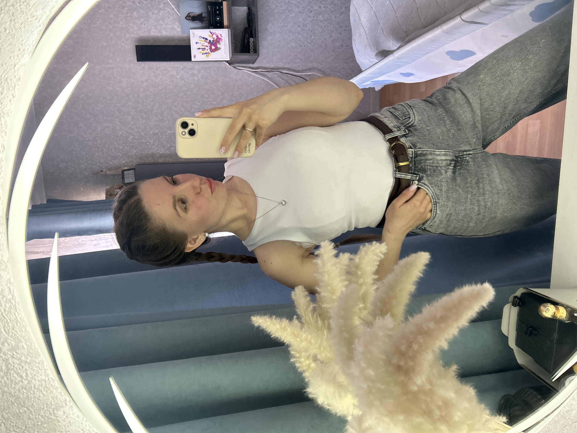 A woman is taking a selfie in a mirror. She is wearing a white t-shirt and grey jeans with a brown belt. Her hair is in a braid. She is holding a cell phone in her right hand. A bouquet of dried flowers is visible in the foreground. In the background is a wall, a shelf with items on it, and a baby crib. The location is Klin, Russia.