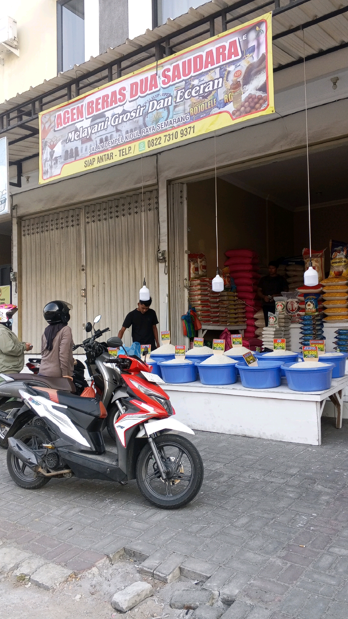 An outdoor scene depicts a rice shop in Semarang, Indonesia. A banner above the storefront reads "AGEN BERAS DUA SAUDARA" and "Melayani Grosir Dan Eceran" (serving wholesale and retail). Below this text, it lists "JALAN DEMPEL KIDUL RAYA SEMARANG" (Dempel Kidul Raya Street, Semarang) and a phone number. The shop has large metal roll-up doors. In front of the shop, rice is displayed in large blue tubs on a white counter. Several sacks of rice are stacked behind the tubs. Two individuals are visible in the scene. One, wearing a black t-shirt and a white cap, stands behind the counter, appearing to be an employee. Another individual, wearing a brown jacket and a black helmet, is seated on a red and white motorcycle parked in front of the shop. A second person, partially visible in a light-colored jacket, stands next to the seated individual. The ground is paved with interlocking bricks.