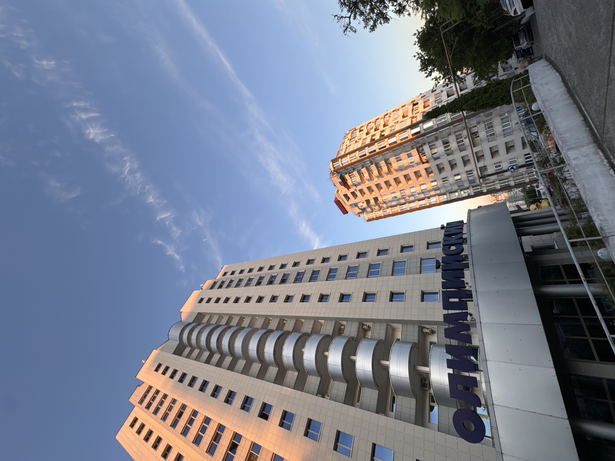 An upward-angled photograph captures two tall buildings against a clear blue sky with wispy white clouds. The time of day appears to be either late afternoon or early morning, as indicated by the warm, golden sunlight illuminating the facades of the buildings.
The prominent building in the foreground, located on the left, is a light-colored high-rise featuring rows of distinctive, rounded, metallic balconies. Numerous rectangular windows are visible across its surface. Vertically arranged large blue letters on its right side spell out "ОЛИМПИЙСКИЙ" (Olimpiyskiy), meaning "Olympic" in Russian.
Behind and to the right of the foreground building stands another multi-story structure, also reflecting the warm sunlight on its windows and upper floors. Bits of green foliage from trees are visible along the right edge of the frame, alongside what appears to be a sidewalk or road with hints of parked vehicles. The sky is largely clear, with only a few delicate, streaky clouds. The low-angle perspective emphasizes the towering height of the urban architecture, set against the peaceful backdrop of the sky. The visible text "ОЛИМПИЙСКИЙ" strongly suggests the location is Sochi, Russia, known for hosting the Olympic Games.