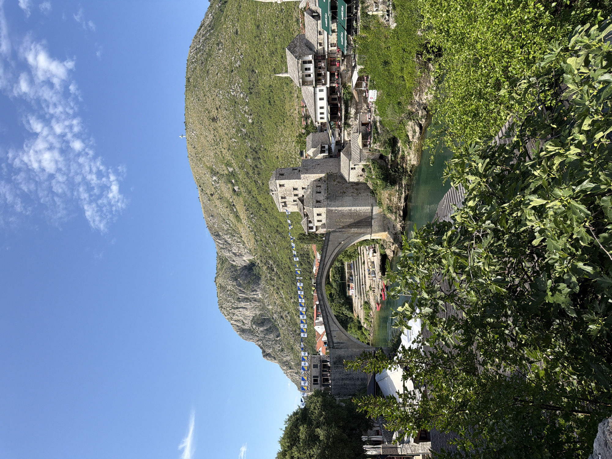 This vertical image captures a panoramic view of the Stari Most (Old Bridge) spanning the Neretva River in Mostar, Bosnia and Herzegovina, under a clear blue sky with scattered white clouds. The foreground features dense green foliage, including trees with fig-like leaves, partially obscuring the river below.
The iconic stone arch bridge dominates the middle ground, connecting the two banks of the river. Above the bridge, a string of small rectangular flags, primarily yellow and blue, is suspended across the river. On the left bank, adjacent to the bridge, are multi-story stone buildings with traditional architecture and reddish-brown tiled roofs. Some structures with white awnings are visible at the base of these buildings. On the right bank, a collection of stone buildings, some with white facades and green awnings (suggesting cafes or restaurants), are built into the rocky incline. A tall, slender stone tower, likely a minaret, is partially visible on the far right. Below the bridge on the right bank, terraced steps lead down to the emerald-green river, where a few red kayaks or small boats are docked. White tent-like structures with text (e.g., "Mostar") are present on the riverbanks.
In the background, two prominent mountains frame the scene. The mountain on the left is rugged and rocky, while the one on the right, Mount Hum, is covered in green scrub and culminates in a prominent white cross at its summit. The sky is bright and largely clear, with a few dispersed cumulus clouds. No individual people are distinctly visible, but the presence of cafes and flags suggests human activity. The overall lighting indicates bright daylight conditions.