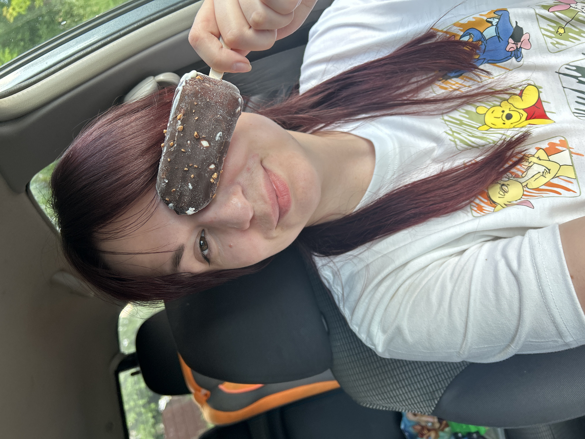 A woman with long, dark red hair is sitting in the driver's seat of a car. She is holding a chocolate-covered ice cream bar with nuts, covering her right eye. She is wearing a white t-shirt with images of Winnie the Pooh characters. The interior of the car is visible, including the car seat and the dashboard. Outside the car, trees and foliage can be seen. The scene is consistent with being in a car parked or driving in a suburban or rural area in Фоки, Russia.