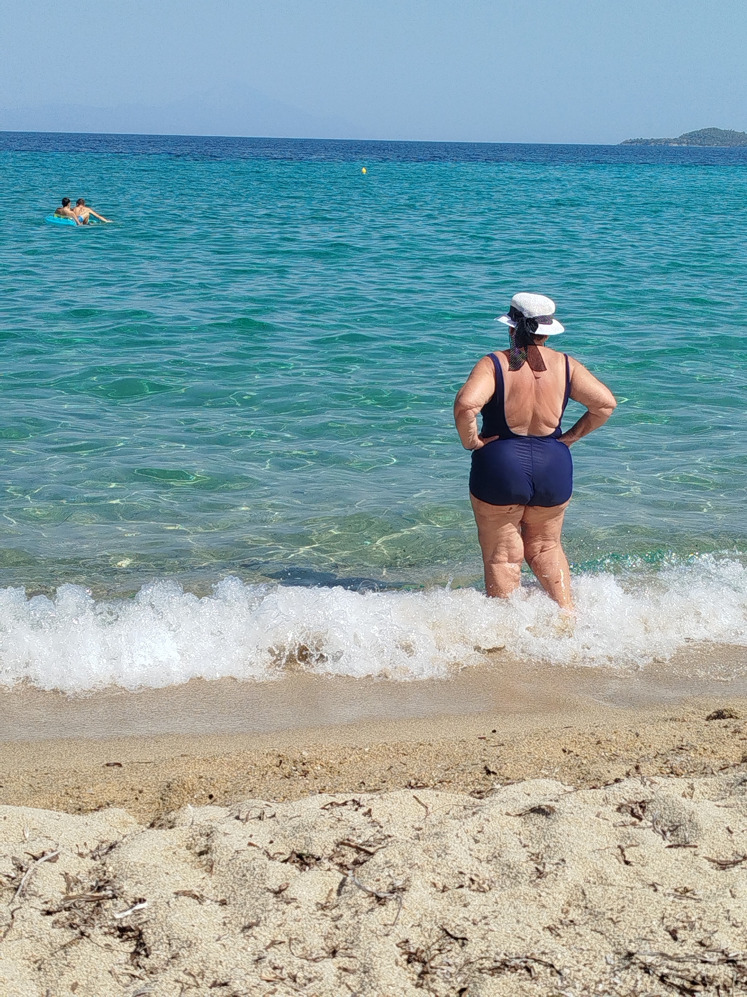 A woman in a navy blue swimsuit and a white sun hat stands with her back to the camera in the shallow waters of a beach. The water is clear and turquoise, with small waves lapping at the shore. Her hands are on her hips. In the background, two people are visible in the water, one on an inflatable ring. The beach is sandy and dotted with dried seaweed. The sky is clear and blue, with a distant hazy coastline. The location is likely a beach in Sithonia, Halkidiki, Greece, known for its clear waters and sandy beaches.