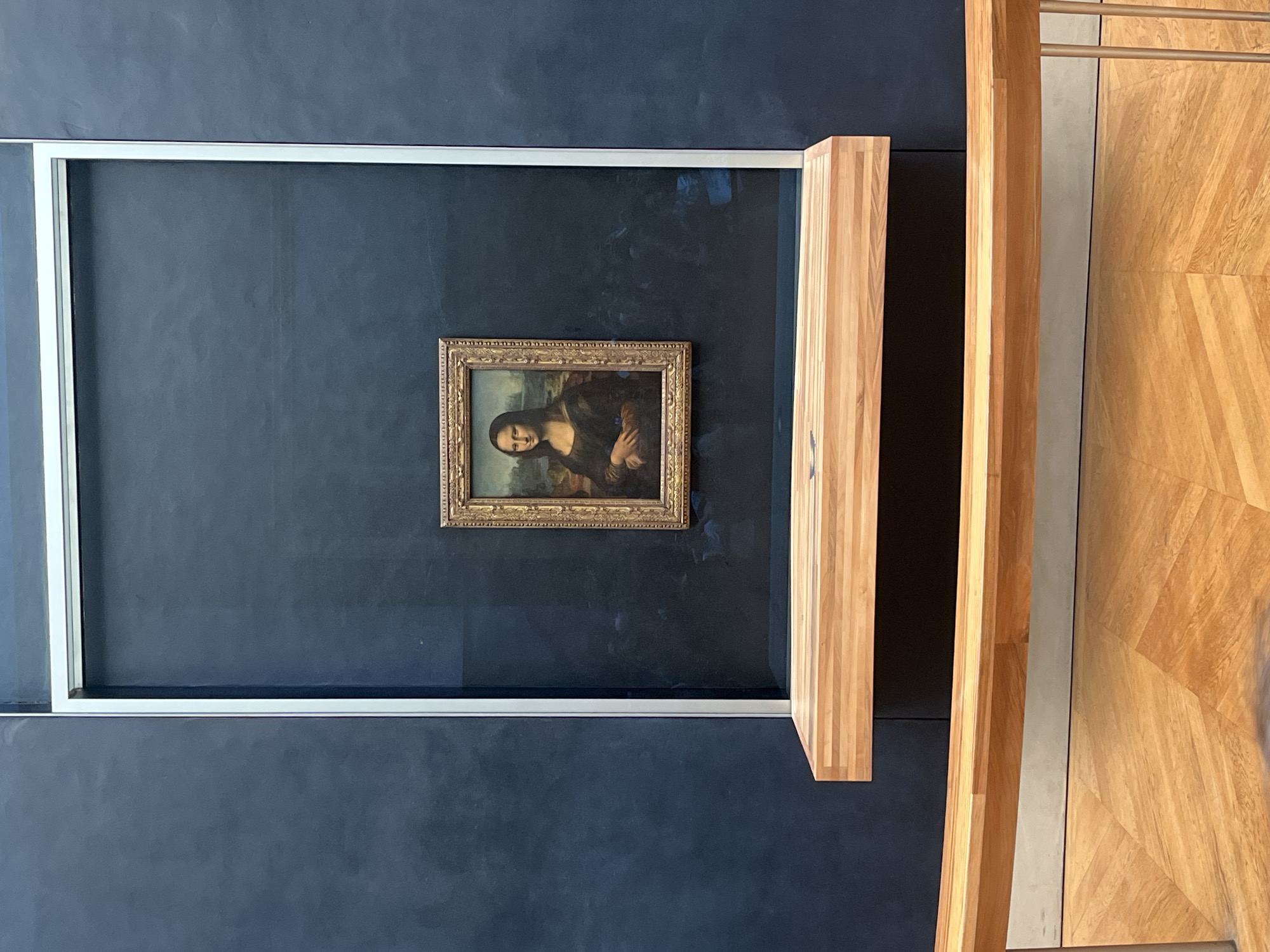 The image displays the iconic Mona Lisa painting by Leonardo da Vinci, housed within a museum setting, specifically the Louvre in Paris, France. The painting, relatively small in size, is centrally positioned against a dark blue-grey wall. It is enclosed within a gold-colored ornate frame and further protected by a large sheet of clear, reflective glass, which forms part of a modern, large white rectangular display frame.
Reflections of faint human figures and ambient light are visible on the protective glass, indicating the presence of visitors viewing the artwork. To the right of the display, there is a prominent, light-colored wooden structure, possibly a barrier or part of the display stand, featuring distinct vertical grain patterns. Further right, another larger wooden structure and a light wooden floor with a geometric parquet pattern (possibly herringbone) are visible, typical of museum flooring. The overall scene suggests a well-maintained and controlled environment designed for public viewing of a prized masterpiece, with elements in place for crowd management, such as the wooden barriers. There is no visible text in the image.
