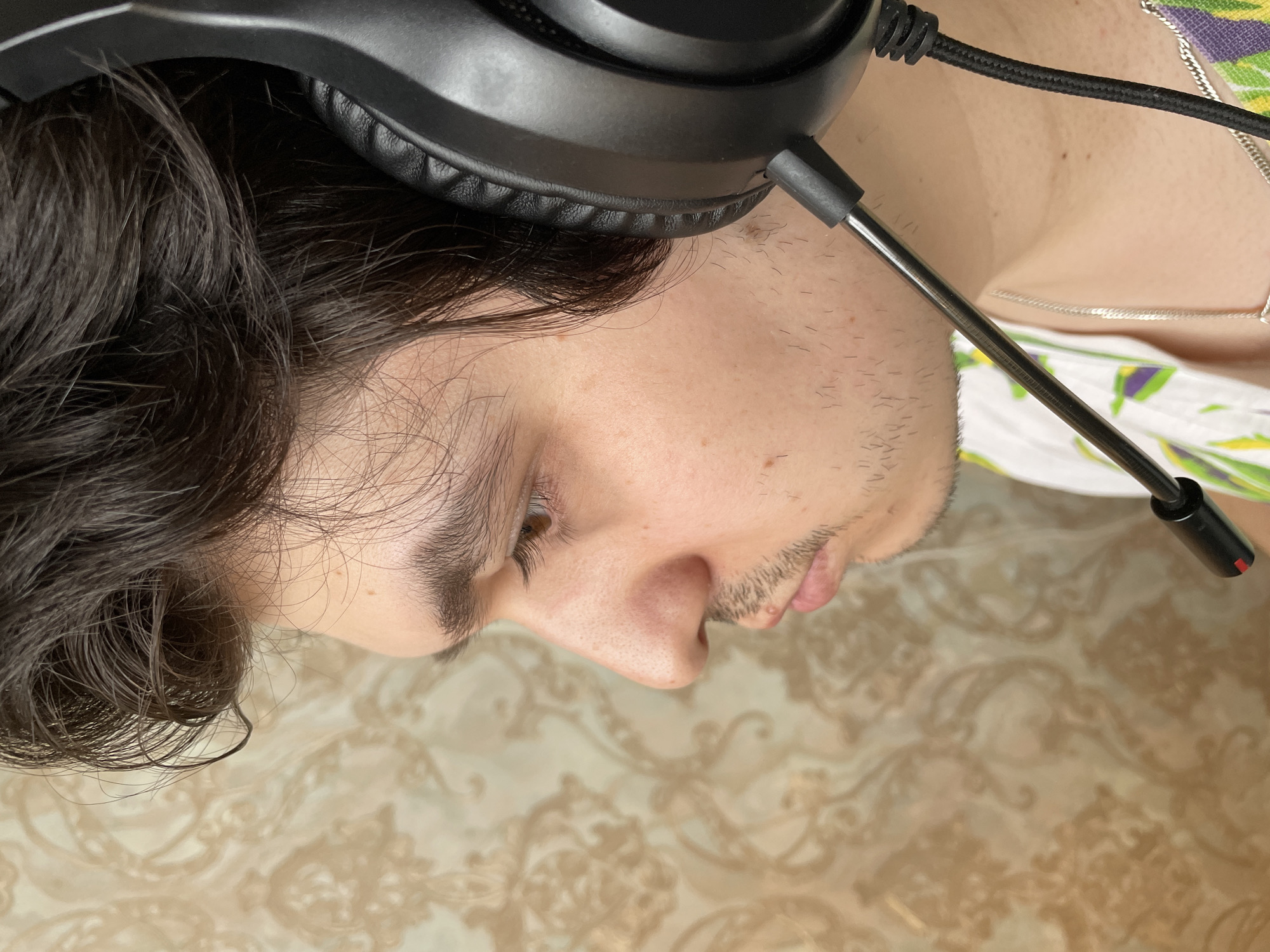 A close-up, low-angle shot of a person wearing black headphones with a boom microphone. The person has dark, wavy hair and is clean-shaven. Their head is tilted downward, and their face is in profile. The background consists of a patterned surface, likely a tablecloth or similar textile. The image does not provide enough information to confirm a location in Izhevsk, Russia.