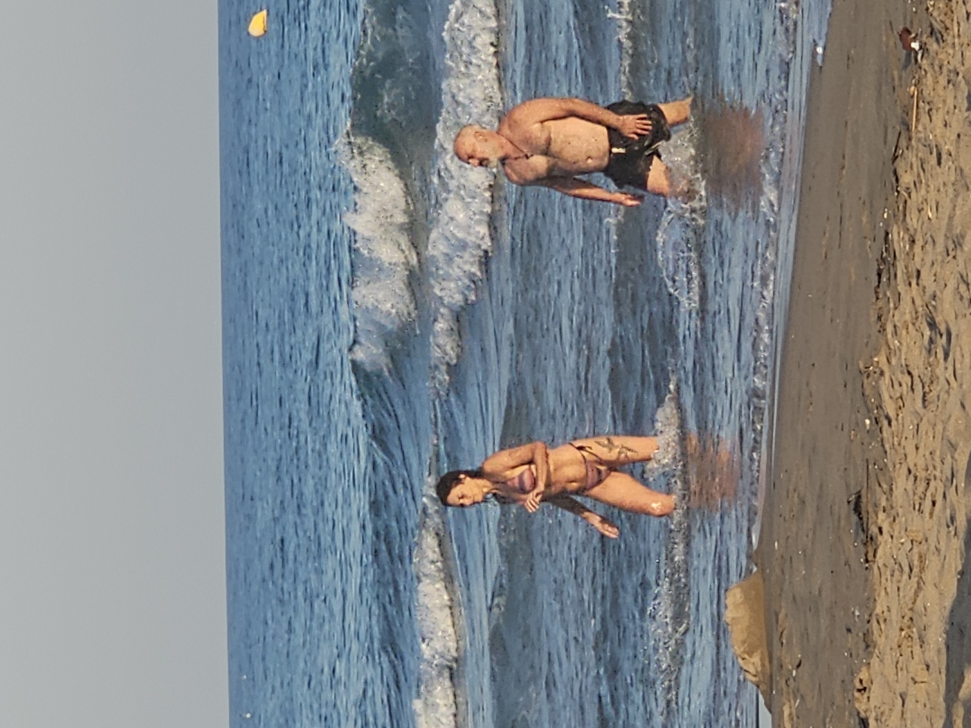 The image depicts two individuals, a woman and a man, wading in the shallow waters of a beach. The woman, on the left, is wearing a patterned bikini and appears to be moving through the water. The man, on the right, is wearing dark swim trunks and has a beard and a necklace. He is also standing in the water. Waves are breaking around them. In the background, the blue sea stretches to the horizon under a pale sky. A yellow buoy is visible in the distance on the right. The foreground shows a sandy beach with some debris. The scene is consistent with a beach in Castelldefels, Spain, known for its long sandy coastline and Mediterranean waters.