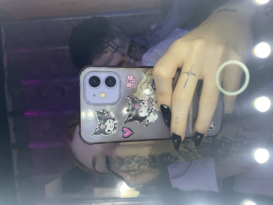 The scene depicts a mirror selfie taken indoors in Buenos Aires, Argentina. A hand, adorned with long, pointed black artificial nails, small black dot tattoos on the fingers, and a black stylized cross tattoo on the ring finger, which also bears a light green ring, holds a light purple smartphone. The phone, an iPhone model with a dual camera system, is encased in a clear cover decorated with two Kuromi stickers, a pink heart, and a pink bow.
Reflected in the mirror are two individuals. A female, presumed to be the individual holding the phone, has light brown hair and is depicted applying makeup to her left eye with an applicator. Her left arm and hand display extensive black tattoos. Partially visible behind her and to her right is a male individual with dark hair and prominent black tattoos covering his forehead and cheek. Both individuals are wearing light-colored shirts. The environment is characterized by dim, purple ambient lighting, with several circular LED vanity lights illuminating the perimeter of the mirror, indicating a dressing area or personal grooming space.