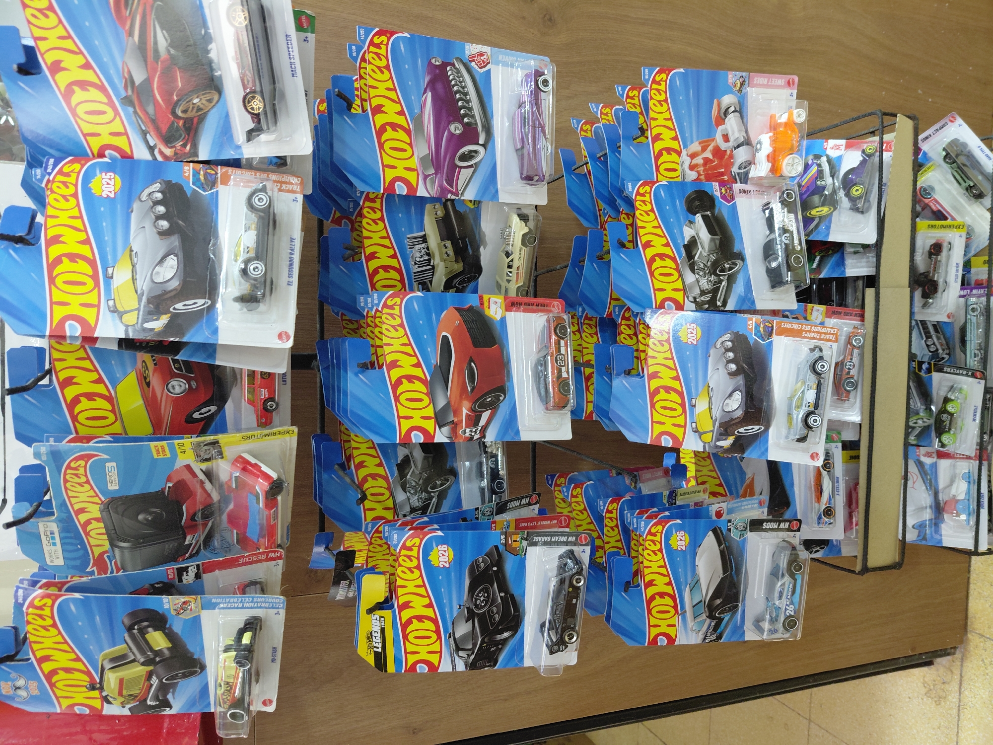 The image shows a close-up, eye-level view of a retail display of Hot Wheels toy cars in their packaging, located in Encarnación, Paraguay. The display is a multi-tiered wire rack with wooden trim, holding numerous blister-packed cars. The cars are predominantly modern, brightly colored, and depicted in dynamic poses on the packaging. The background consists of a light-colored wall and a portion of a wooden display support. The lighting is even, suggesting an indoor retail environment. There are no people or visible interactions in the image. The focus is solely on the product presentation.