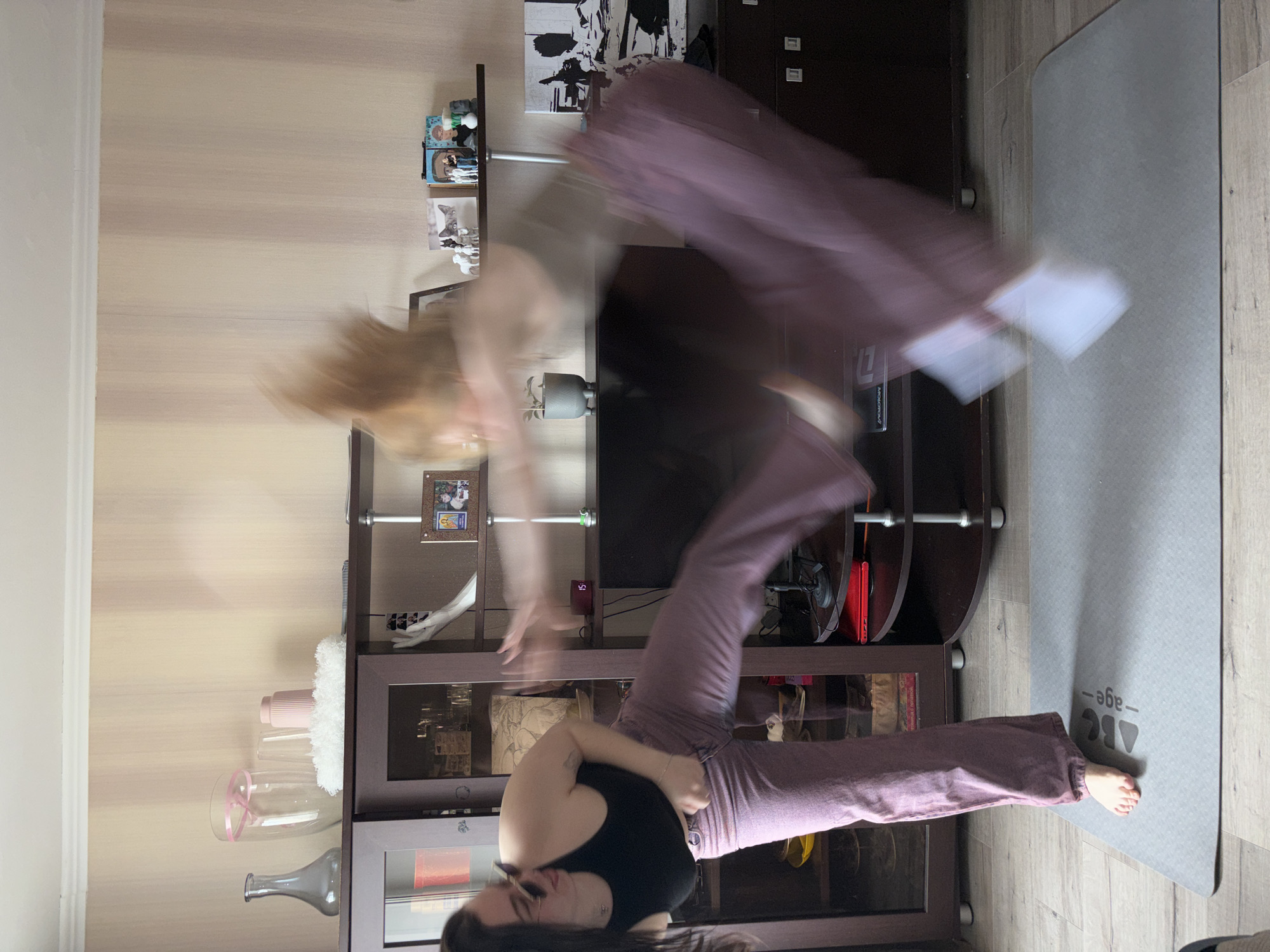 A blurred image shows a person indoors, possibly in an apartment. The location is suggested to be Perm, Russia, based on the prompt.
The person is in motion, with evidence of a double exposure or rapid movement during the image capture. The subject appears to be performing some form of exercise or dance movement. They are wearing a black tank top and maroon or mauve pants.
The environment includes a wooden entertainment center with shelves and glass cabinets containing decorative objects, vases, and possibly books. There is a gray yoga mat on the floor. A shelf above the entertainment center holds pictures and decorative items. The walls are a light beige color.
