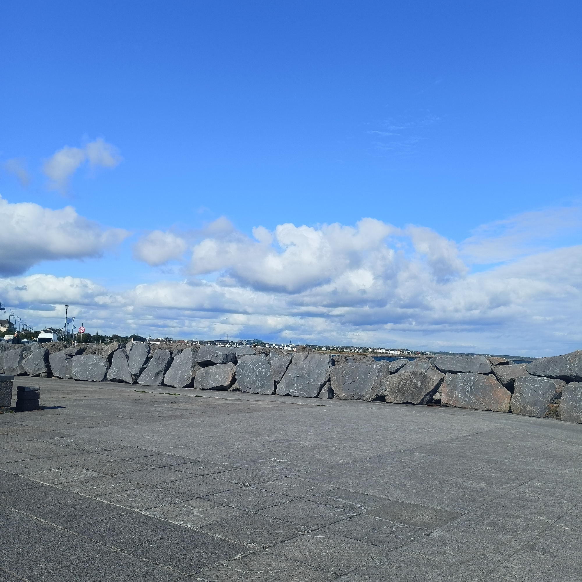 The image shows a view of the Salthill Promenade in Galway, Ireland. In the foreground, there is a paved area made of grey interlocking stones, showing signs of wear and weathering. Beyond the paved area, a large, rough stone wall or barrier is constructed from massive, grey boulders of varying sizes and shapes. This wall runs horizontally across the mid-ground of the image. In the background, a distant townscape is visible, with buildings and structures suggesting a coastal settlement. Above the town, a bright blue sky is dotted with fluffy white clouds, indicating fair weather. The overall impression is a scene of coastal infrastructure, likely serving as a breakwater or promenade wall, with a clear sky and a glimpse of a town on the horizon. There are no people visible in the image.