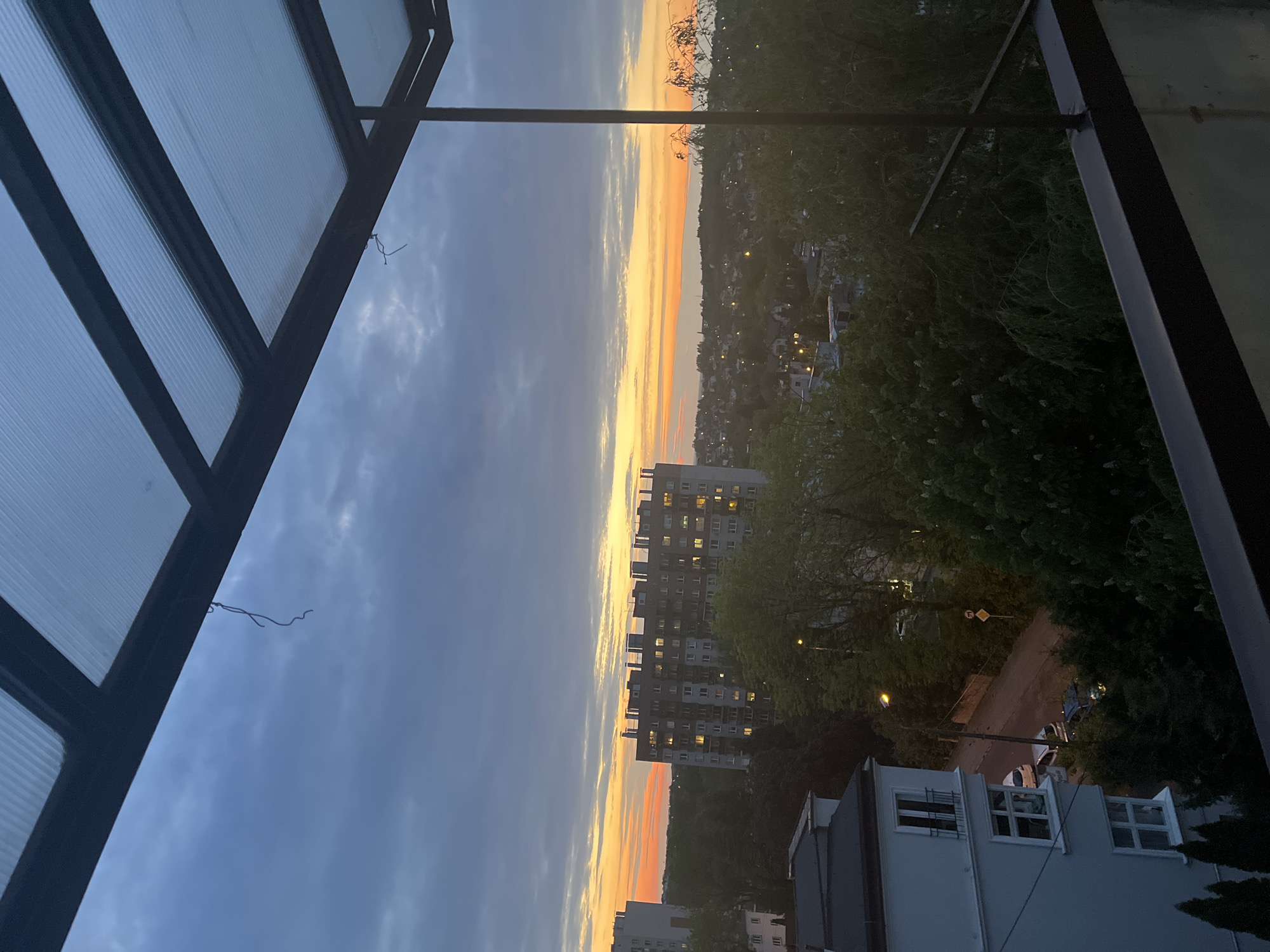 The image depicts a cityscape at dusk or dawn, likely taken from a high vantage point such as a balcony or rooftop. The sky transitions from orange to blue, indicating the transition from day to night or vice-versa. Buildings are visible in the distance, including a tall apartment building with many illuminated windows, suggesting residential occupancy. Streetlights illuminate a road lined with trees. In the foreground, a metallic or dark-colored structure frames the view on the left side, partially obscuring the sky. A white building is located at the bottom right corner. The location appears to be Unknown, Unknown.