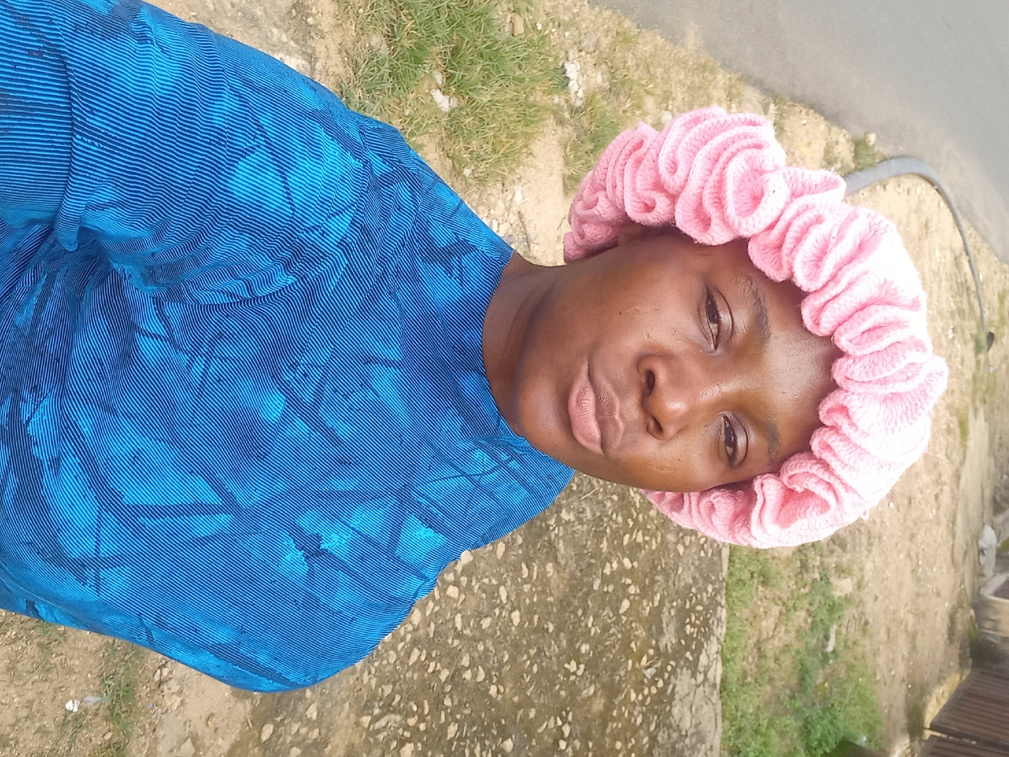 A young Black woman is captured in a close-up, outdoor, selfie-style photograph. She is wearing a pink knitted bonnet with ruffled detailing around the edge and a blue garment with a striped and patterned design. Her face is turned slightly towards the camera, with her lips pursed. The background suggests an urban or semi-urban environment in Jos, Nigeria, with a paved road or sidewalk on the left and a dirt and grassy area on the right, possibly a residential or commercial space with a fence visible in the distance. The lighting indicates it is daytime.